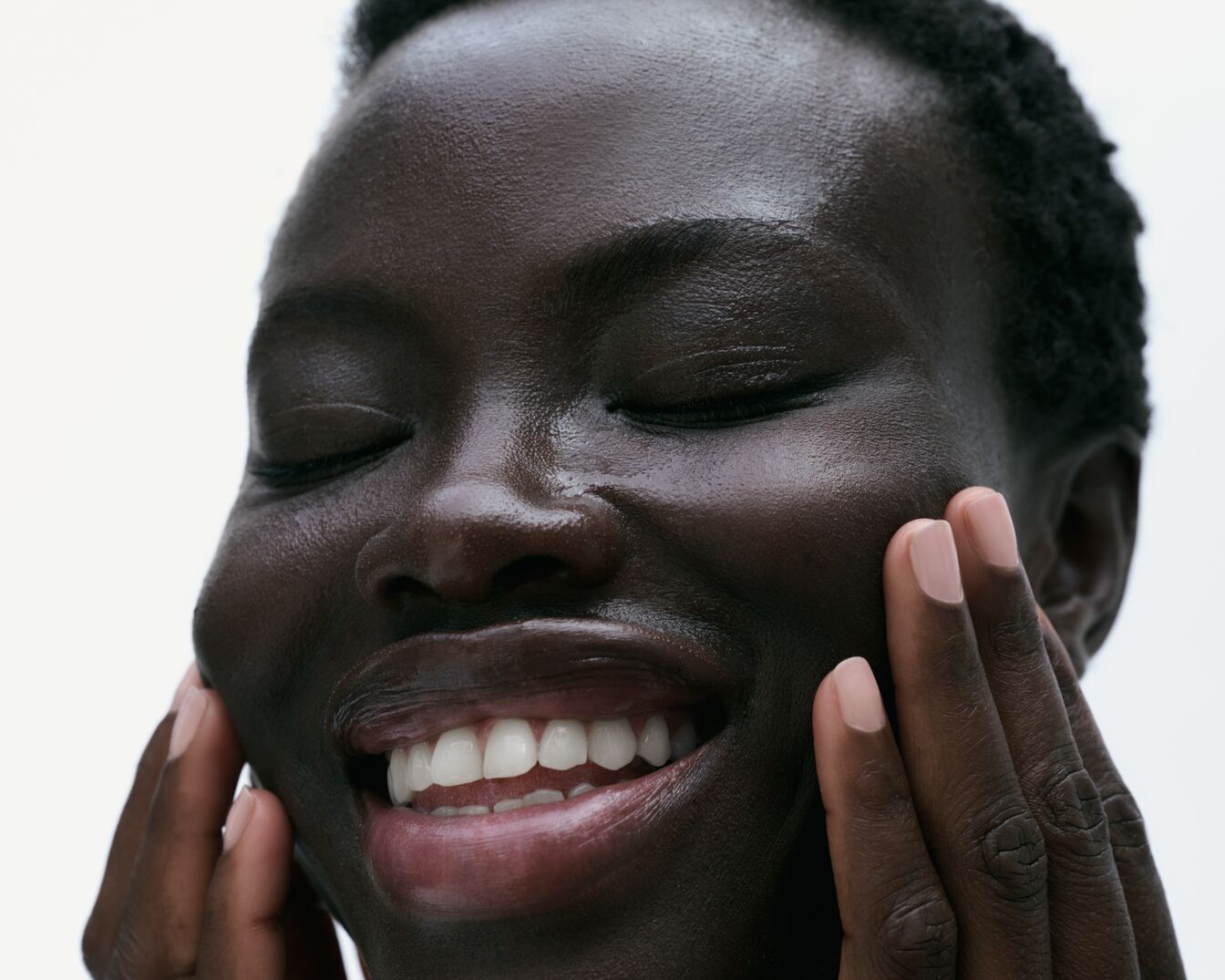 Nahaufnahme einer strahlenden, dunkelhäutigen Frau mit geschlossenen Augen und einem breiten Lächeln, die ihre glatte, glänzende Haut berührt.