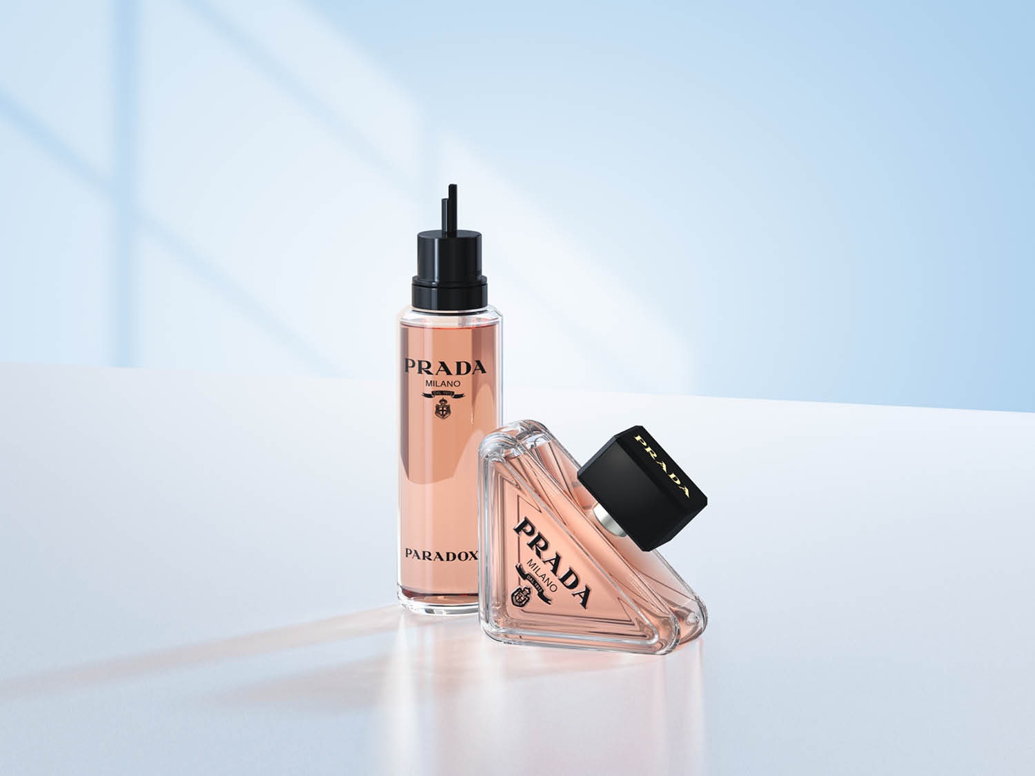 Twee flessen Prada Paradox geur, één lang en rechtopstaand, de andere driehoekig en liggend, beide gevuld met een roze vloeistof.