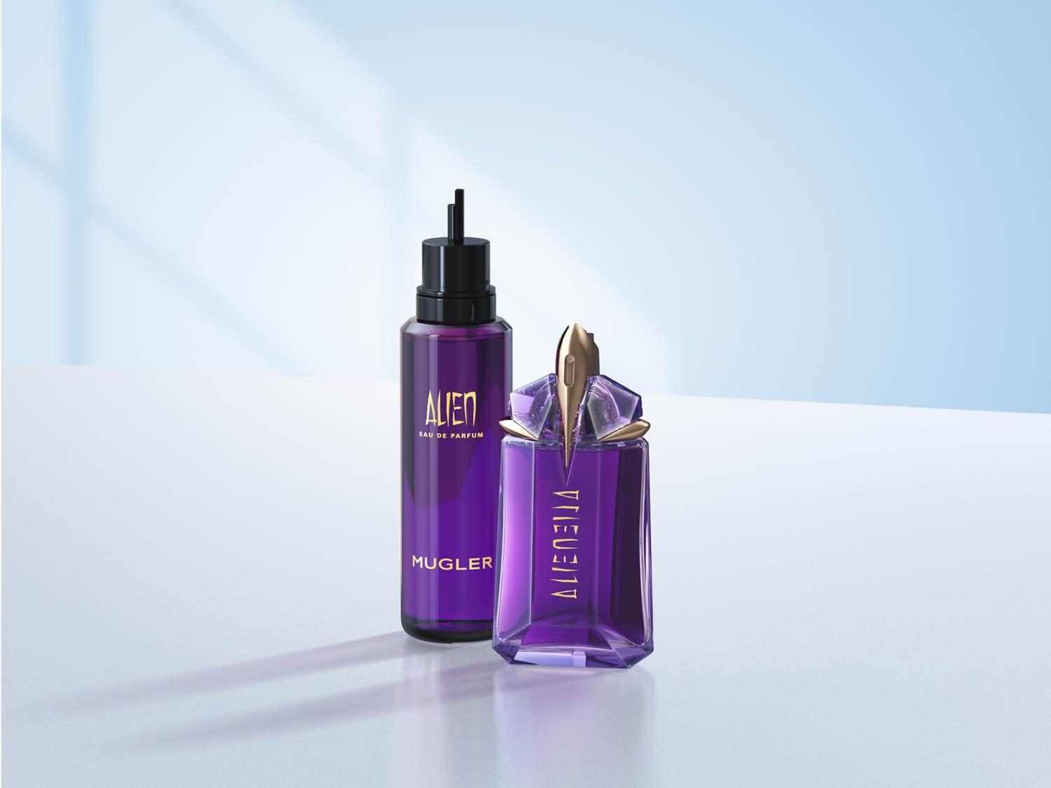 Twee flessen Alien Eau de Parfum van Mugler, de ene is een navulling met een zwarte dop en de andere is een fles in de vorm van een paarse edelsteen met gouden accenten.