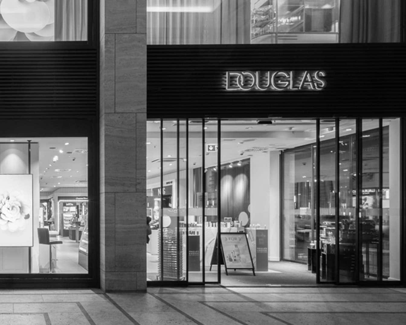 Juodai baltas Douglas parduotuvės fasadas su stiklinėmis durimis ir langu, viršuje užrašas „DOUGLAS“.