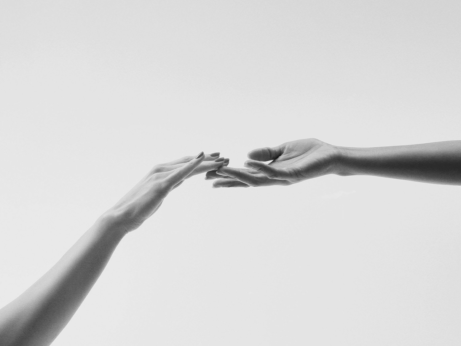 Juodai baltame fone matyti dvi rankos, viena su raudonu manikiūru, siekiančios viena kitos.