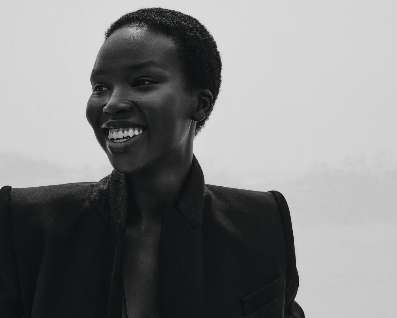 Schwarz-Weiß-Porträt einer lächelnden, dunkelhäutigen Frau mit kurzen, dunklen Haaren, die einen dunklen Blazer trägt und zur Seite schaut.