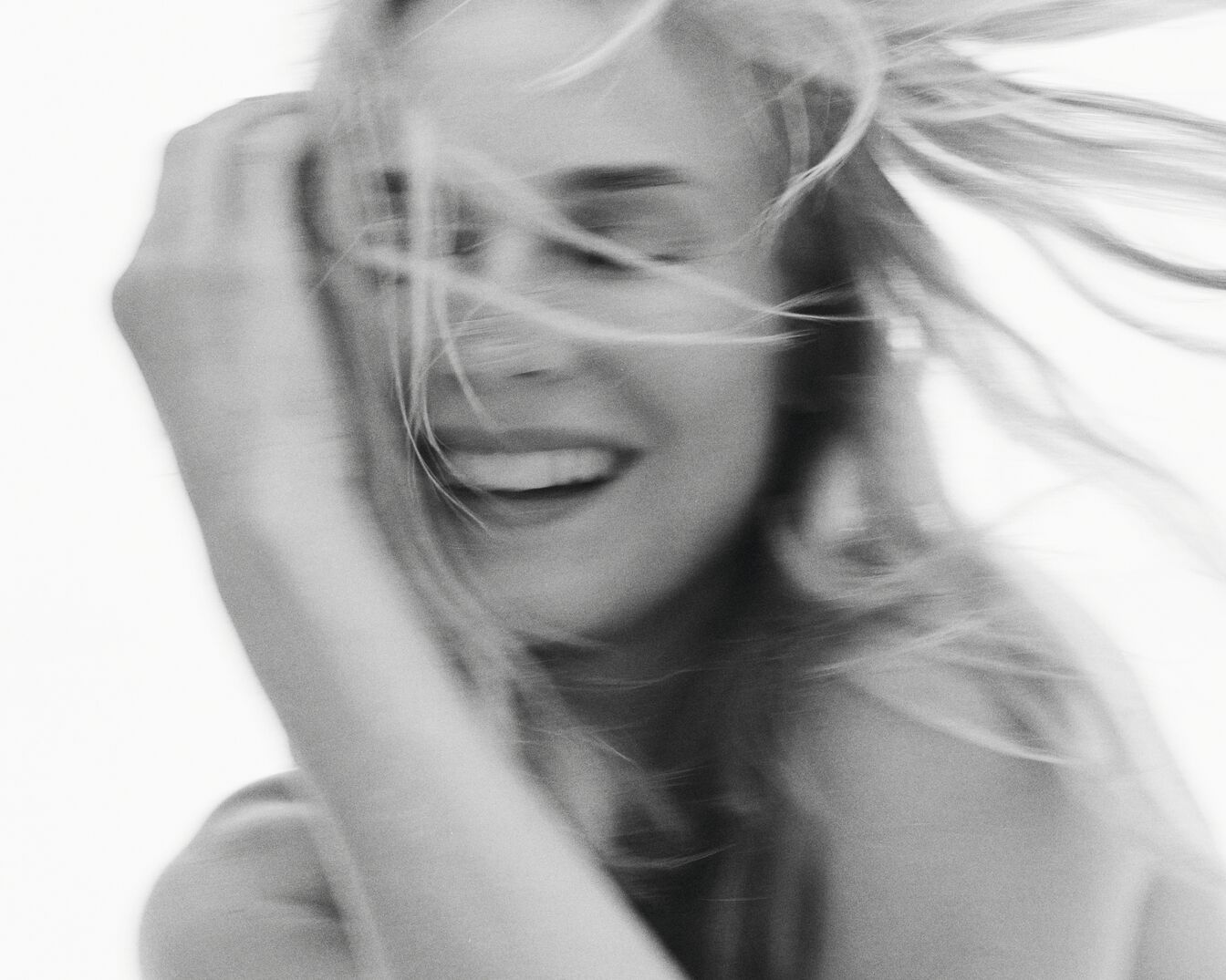 Nahaufnahme einer lachenden Frau mit blonden, im Wind wehenden Haaren, die ihre Hand ins Haar legt, in Schwarzweiß.