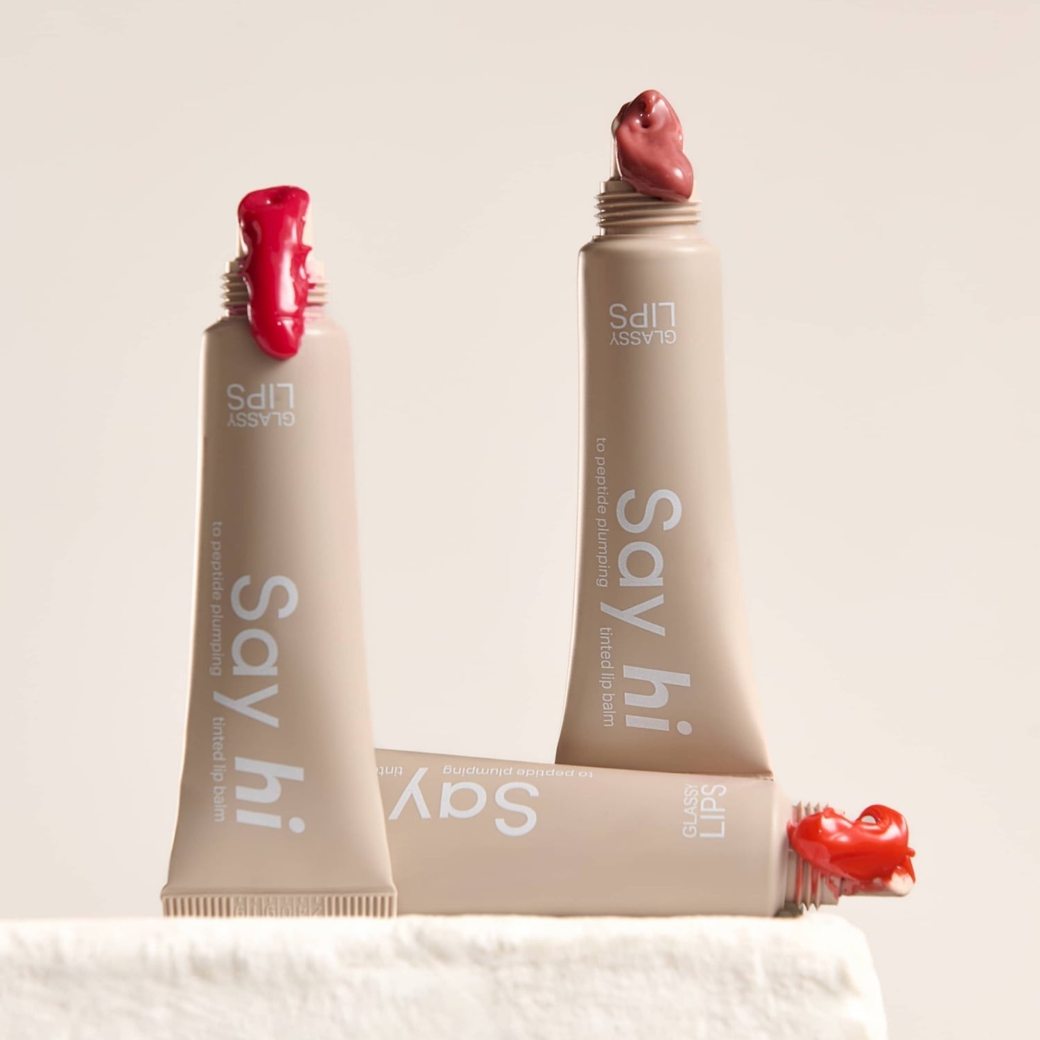 Trzy tubki balsamu do ust Glassy Lips w odcieniach czerwieni i różu, z napisem „Say hi to peptide plumping tinted lip balm”.