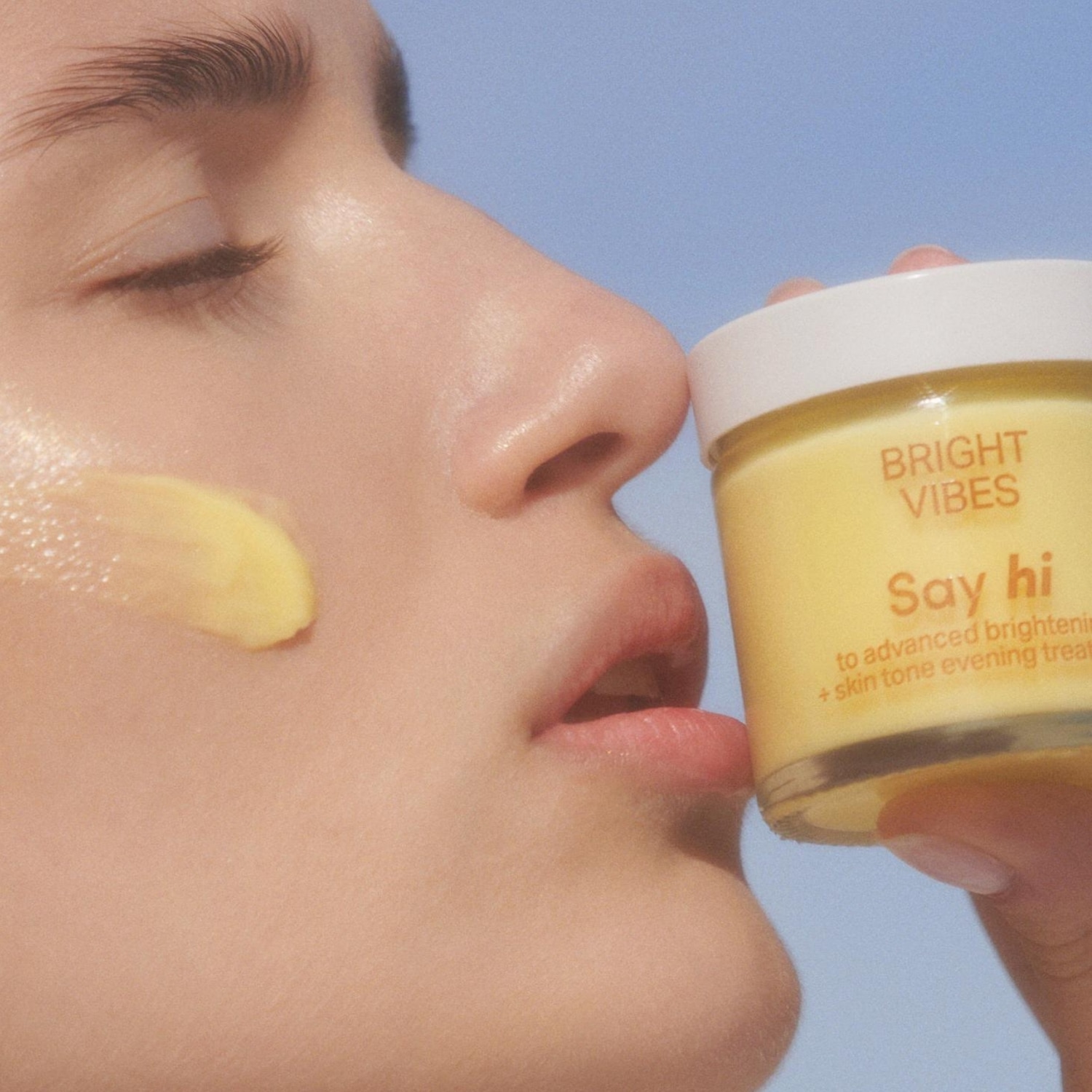 Kobieta nakłada na twarz krem Bright Vibes o żółtej konsystencji z napisem „Say hi to advanced brightening + skin tone evening treatment”, aby uzyskać efekt rozświetlonej i wyrównanej cery.