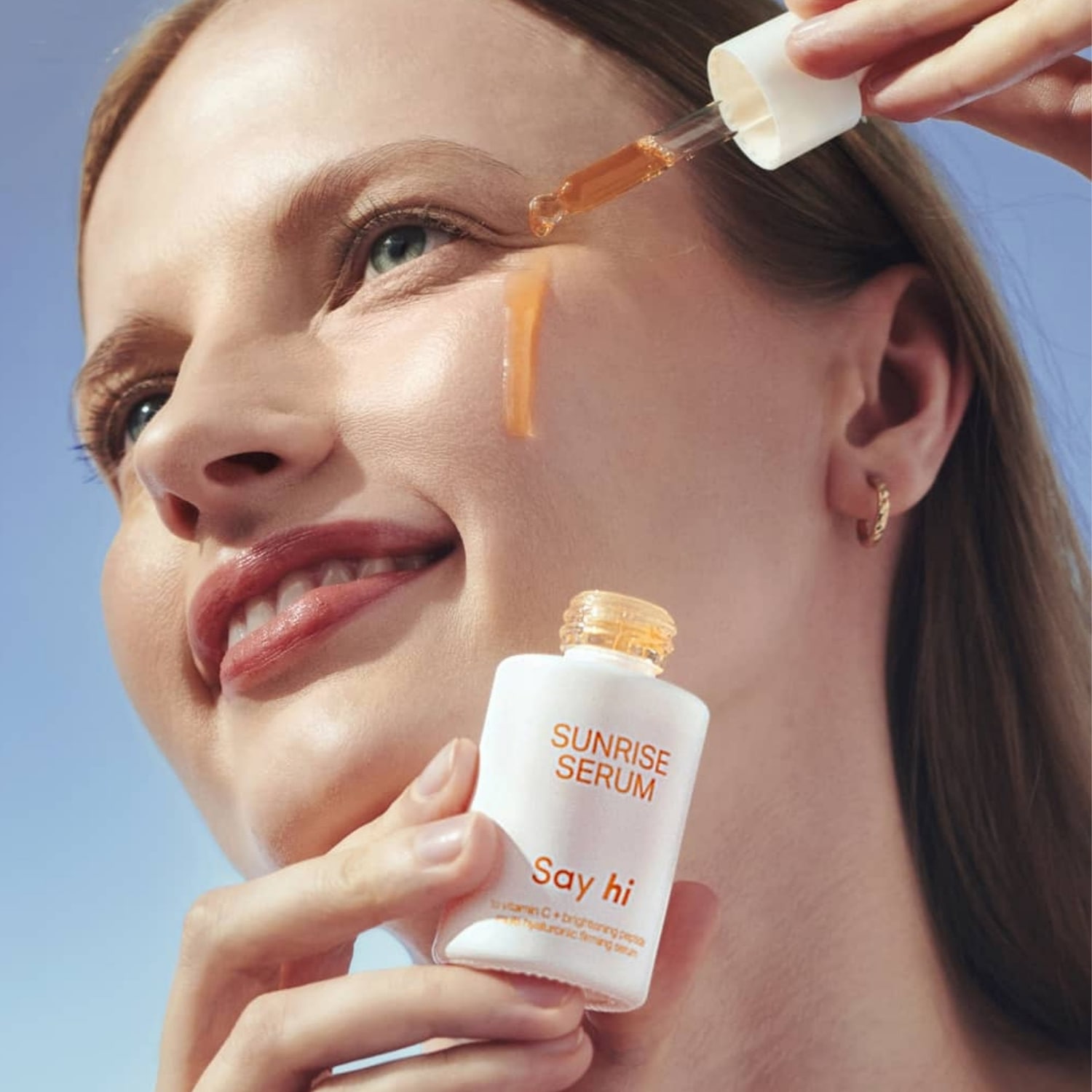 Uśmiechnięta kobieta aplikuje serum z witaminą C marki Say hi na twarz za pomocą pipety, trzymając białą butelkę z napisem Sunrise Serum.
