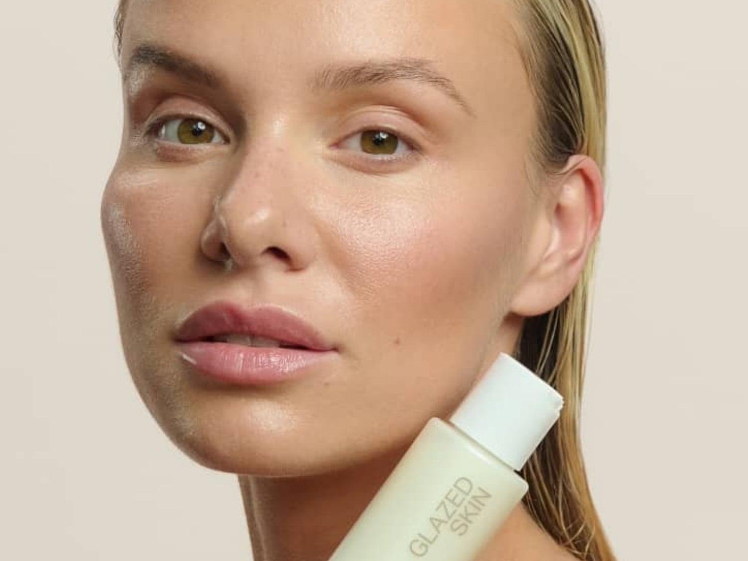 Blond modelka z zielonymi oczami trzyma butelkę produktu do pielęgnacji skóry Glazed Skin, prezentując efekt promiennej i nawilżonej cery.