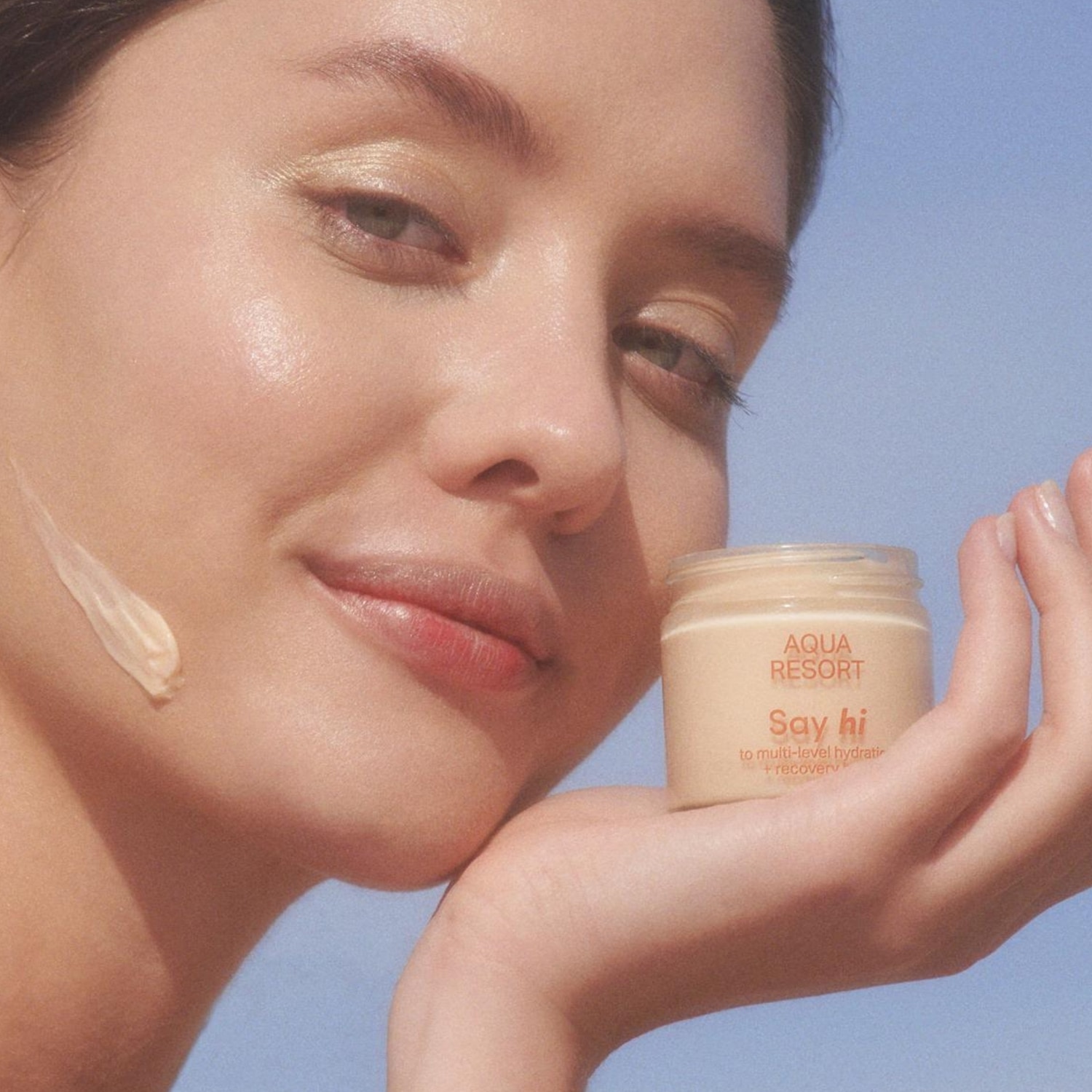 Kobieta z kroplą kremu na policzku prezentuje krem Aqua Resort Say Hi, który zapewnia wielopoziomowe nawilżenie i regenerację.