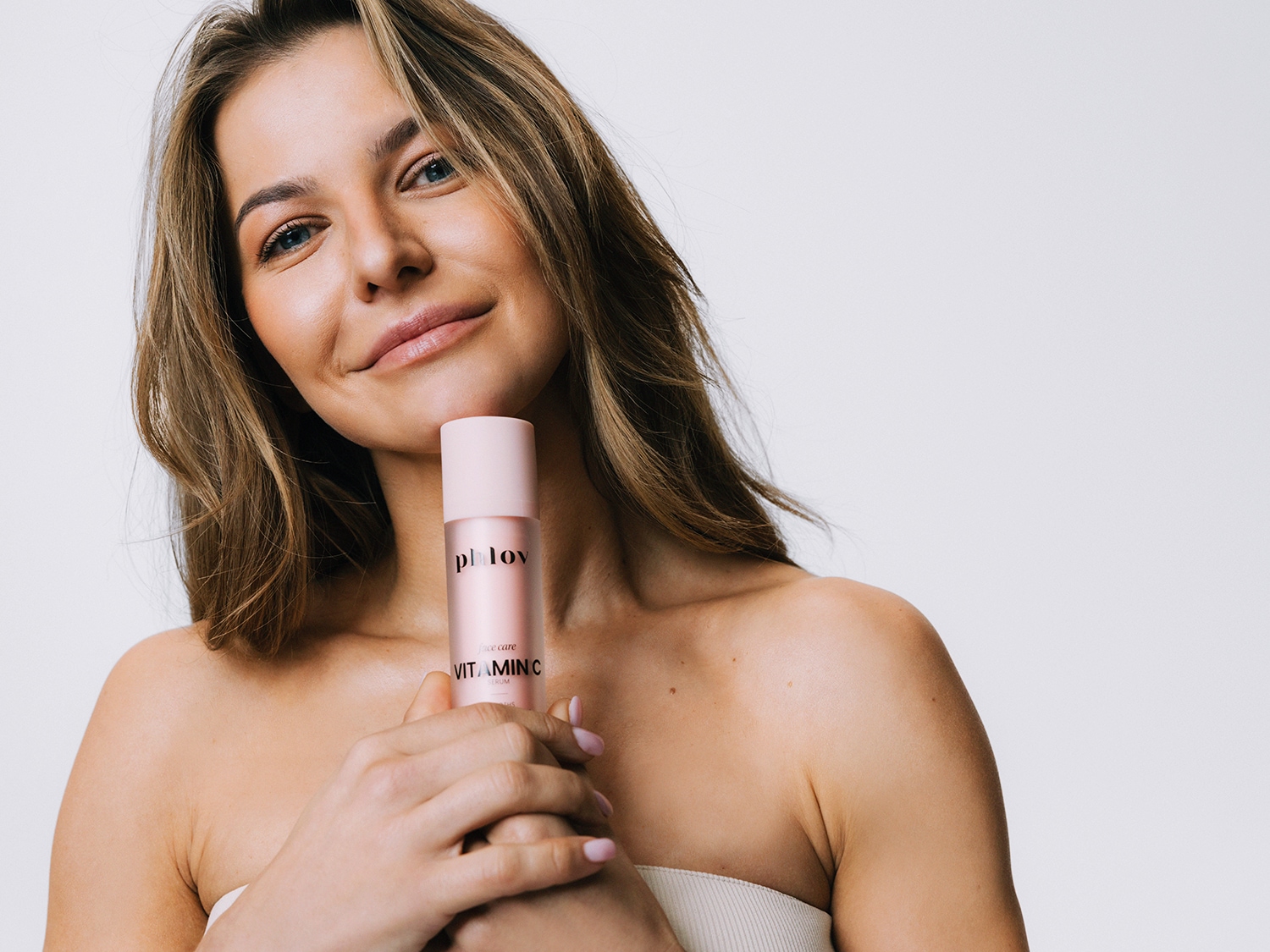Uśmiechnięta kobieta trzyma w dłoniach różową butelkę serum do twarzy Phlov Vitamin C, prezentując produkt do pielęgnacji skóry.