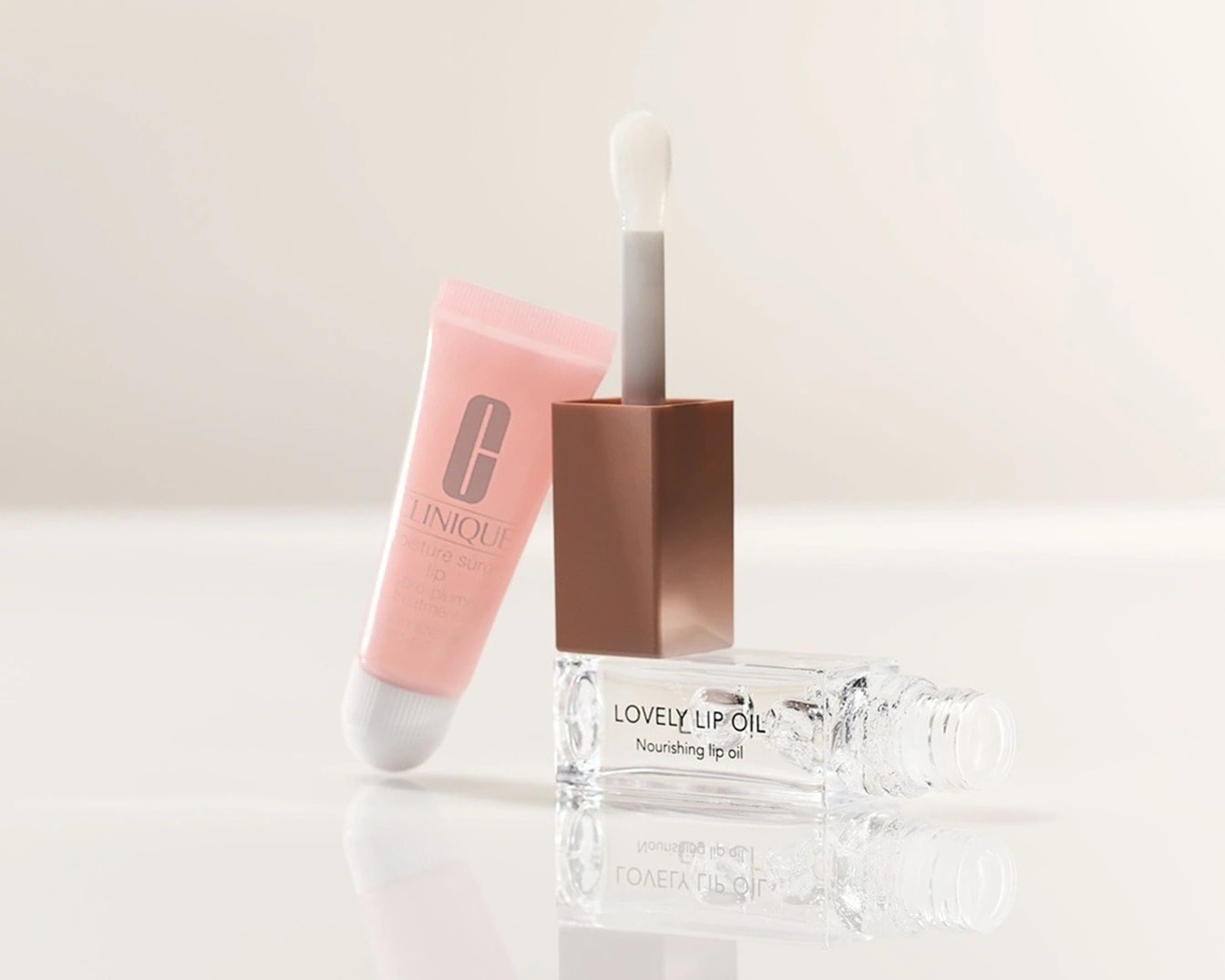 Švelnus rožinis Clinique lūpų blizgesys, Lovely Lip Oil maitinamasis lūpų aliejus skaidriame buteliuke ir aplikatorius su rudu dangteliu ant blizgaus paviršiaus.