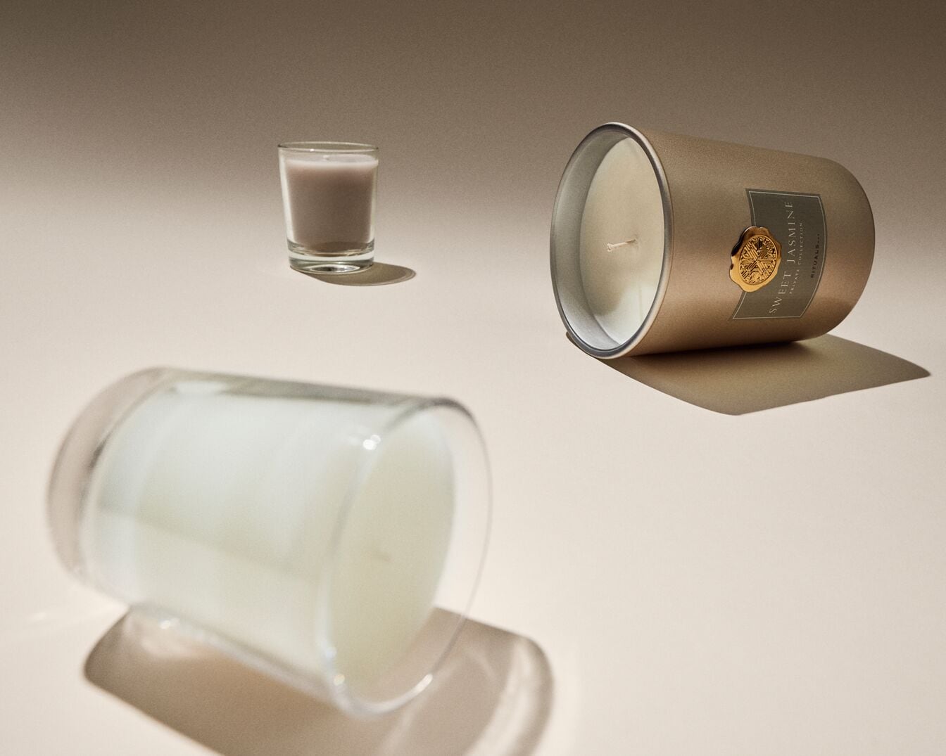 Trys žvakės: maža stiklinė žvakė, didelė žvakė stikliniame inde ir auksinė žvakė su etikete „Sweet Jasmine Rituals Private Collection“.