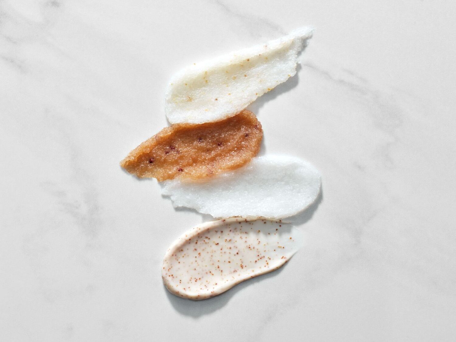 Keturių skirtingų spalvų ir tekstūrų šveitiklių tepinėliai ant balto marmurinio paviršiaus.