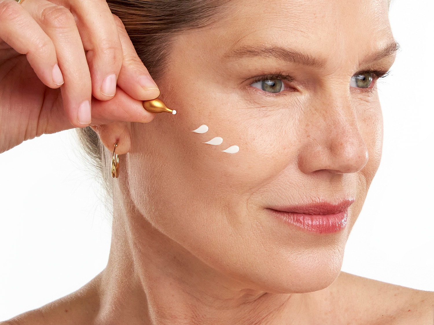 Una mujer aplica un suero facial blanco en su rostro desde una cápsula dorada para hidratar y rejuvenecer la piel.