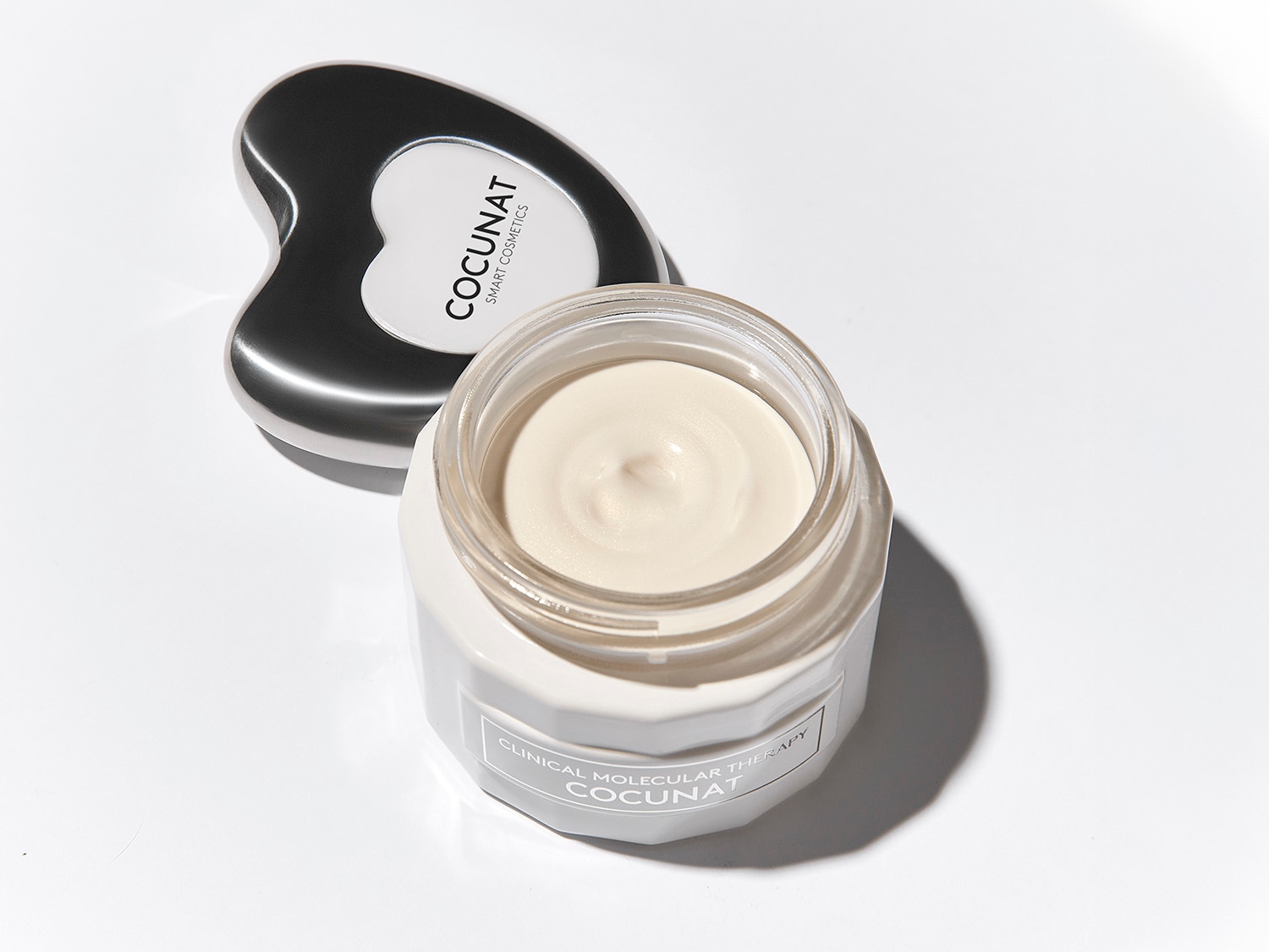 Aquí se muestra un tarro de crema facial Cocunat junto a una herramienta de masaje facial negra y plateada, ambos sobre un fondo blanco brillante.