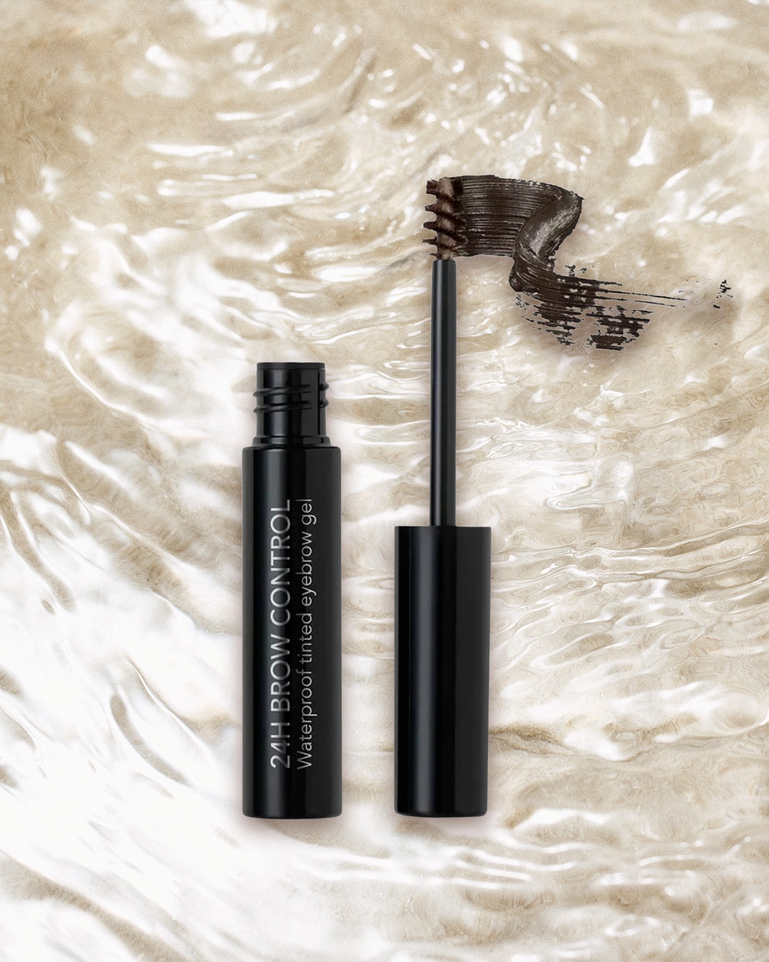 Mascara Benefit in confezione argentata con gocce d'acqua, accanto allo scovolino nero, su sfondo sabbioso e roccia chiara.