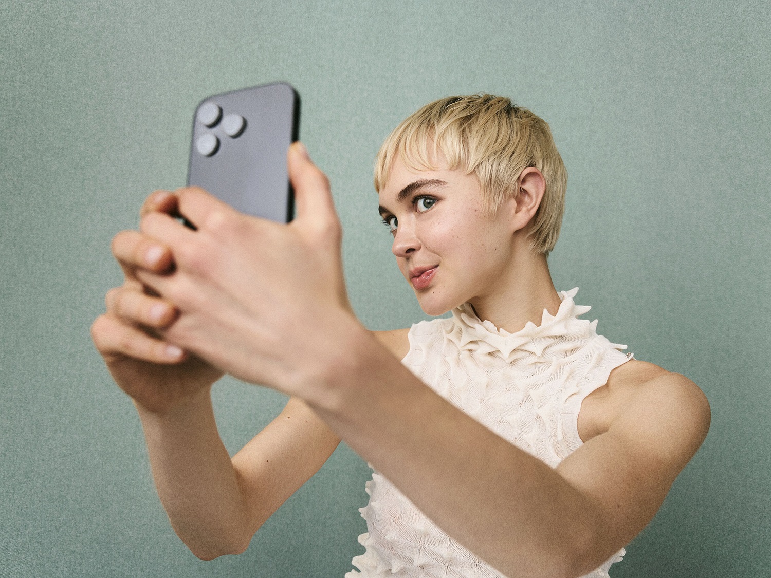 Eine junge Frau mit blonden, kurzen Haaren macht ein Selfie mit ihrem schwarzen Smartphone vor einem hellgrünen Hintergrund.