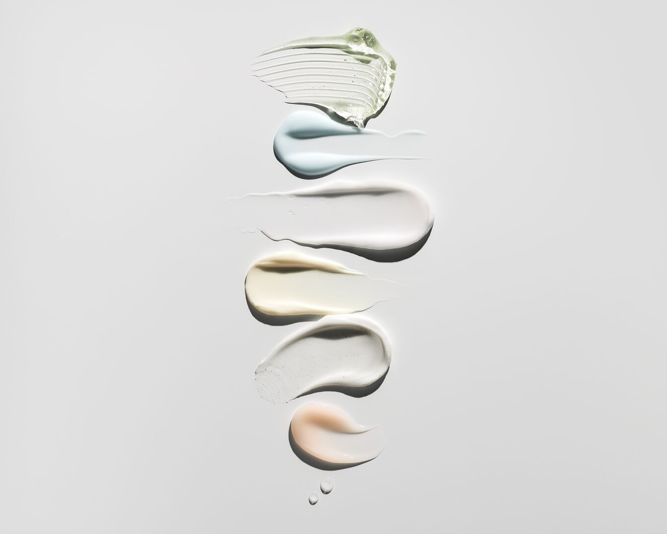 Šešios skirtingų spalvų ir tekstūrų kosmetikos priemonės, įskaitant žalią gelį, mėlyną kremą, baltą losjoną, gelsvą serumą, pilką šveitiklį ir persikinį balzamą.