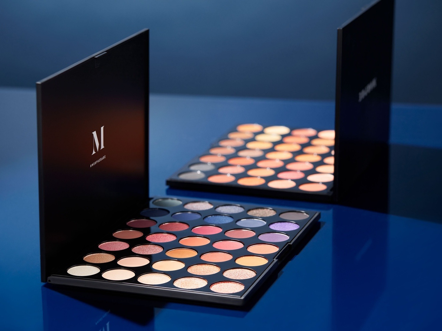 Twee Morphe oogschaduwpaletten met een verscheidenheid aan kleuren, van neutrale tinten tot levendige blauw- en paarstinten, gepresenteerd in strakke, zwarte verpakkingen.