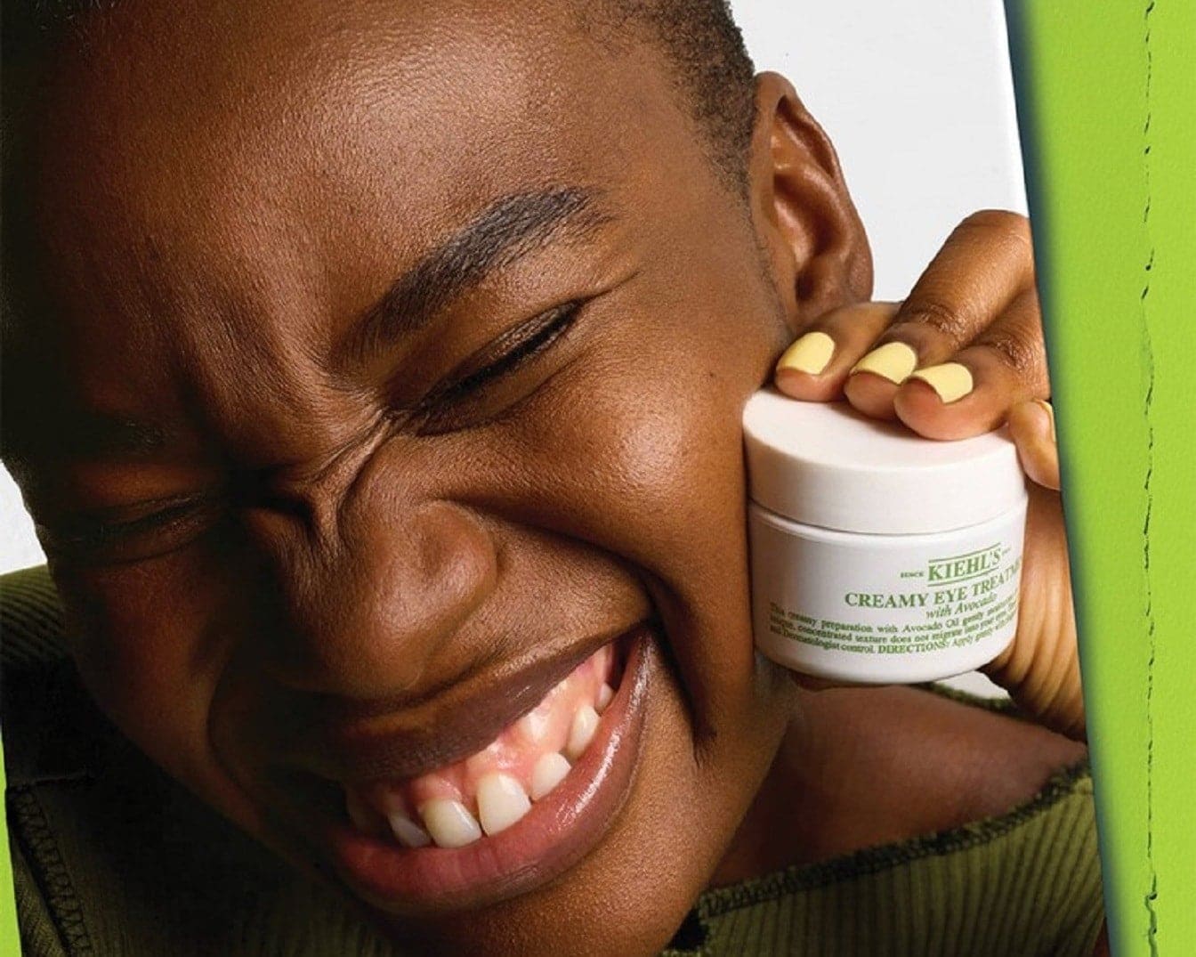 Uśmiechnięta kobieta trzyma krem pod oczy Kiehl's Creamy Eye Treatment z awokado w białym słoiczku, prezentując jego bogatą konsystencję.