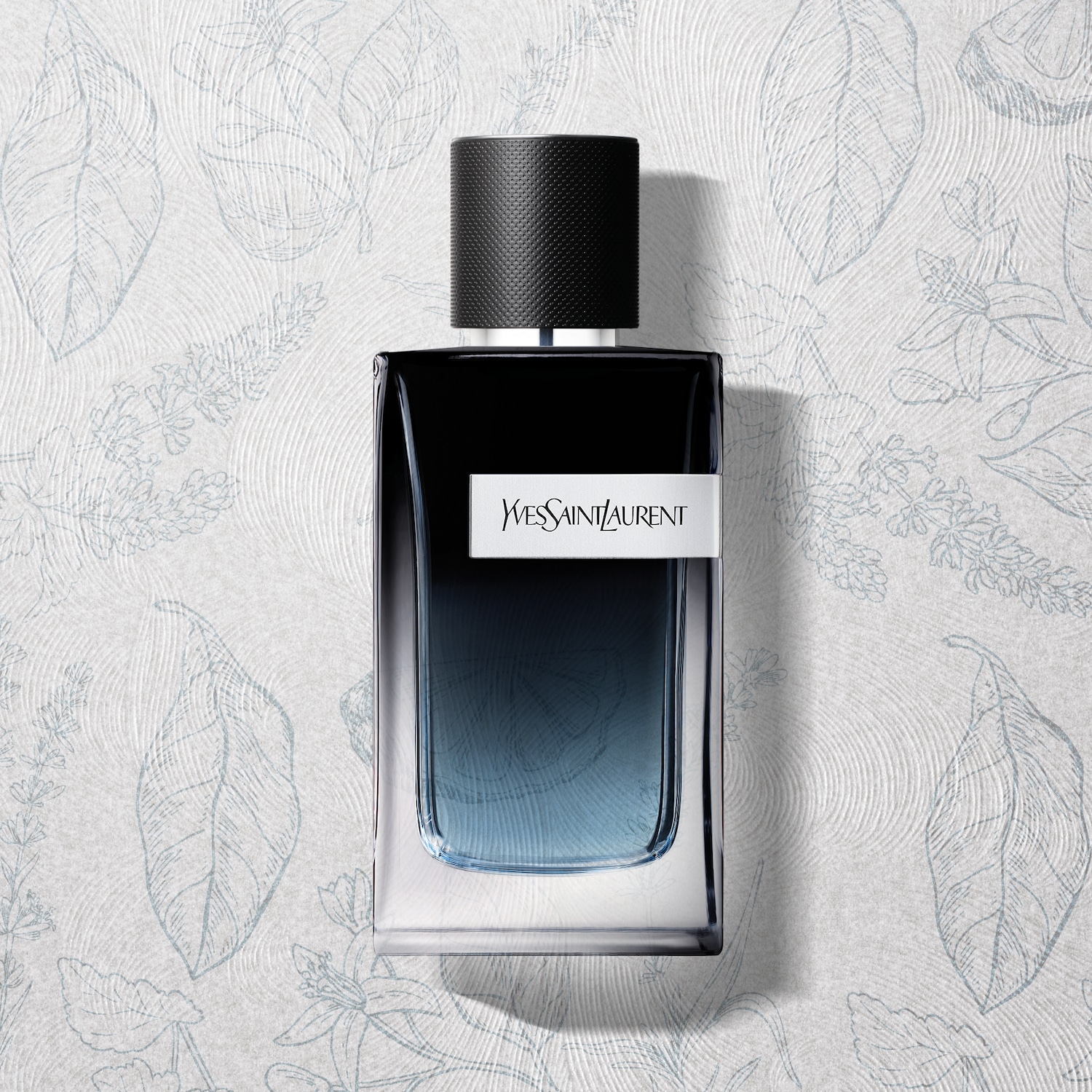 Butelka perfum Yves Saint Laurent w czarnym flakonie z gradientem przechodzącym w błękit, na tle z motywem liści i kwiatów.