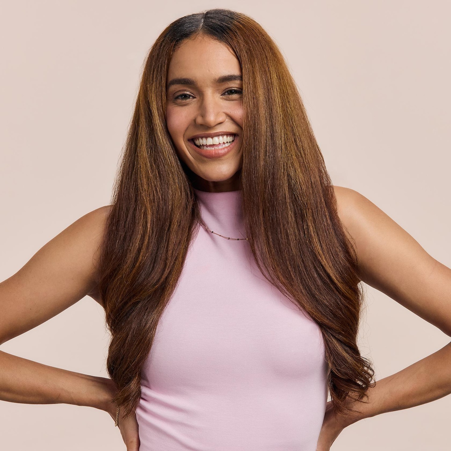 Egy mosolygós, hosszú, barna hajú nő rózsaszín felsőben, kezeit csípőre téve, boldogan néz.