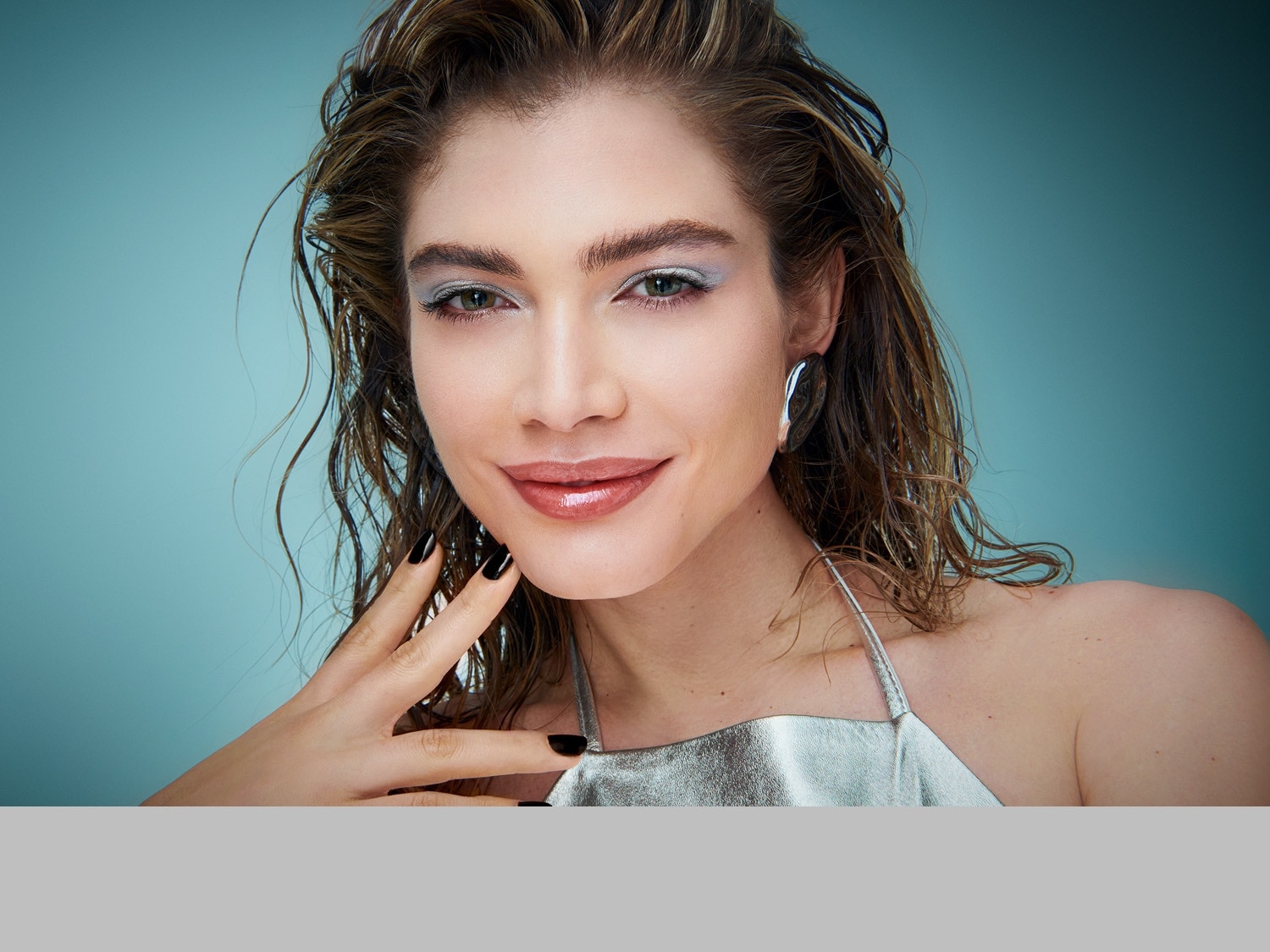 Nahaufnahme einer Frau mit glänzendem, nassem Haar, silbernem Top und passendem Lidschatten, die mit lackierten Nägeln ihr Gesicht berührt.