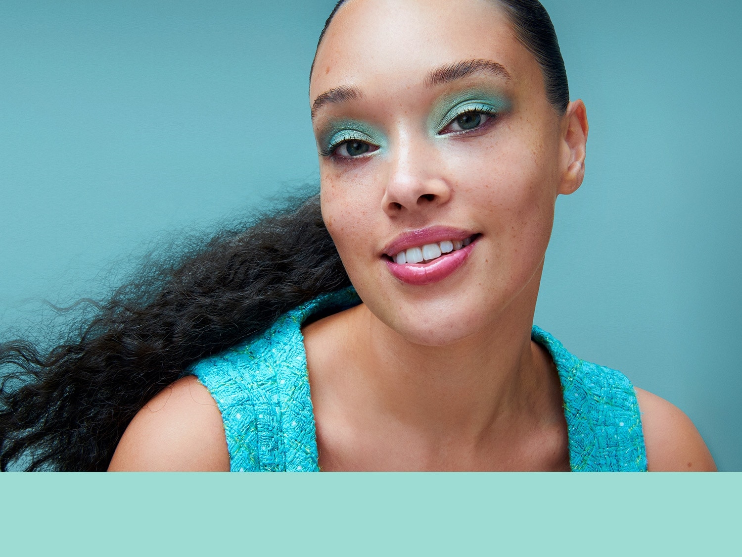 Nahaufnahme einer Frau mit hellgrünem Lidschatten, passendem Oberteil und dunklen, lockigen Haaren vor einem türkisfarbenen Hintergrund.