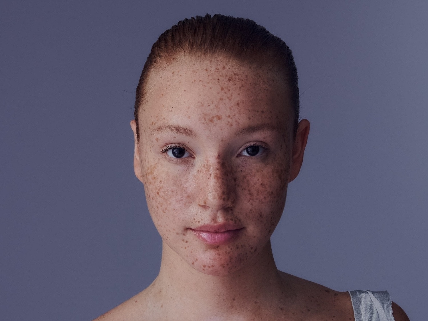 Porträt einer jungen Frau mit Sommersprossen und zurückgekämmten roten Haaren vor einem schlichten, hellvioletten Hintergrund.
