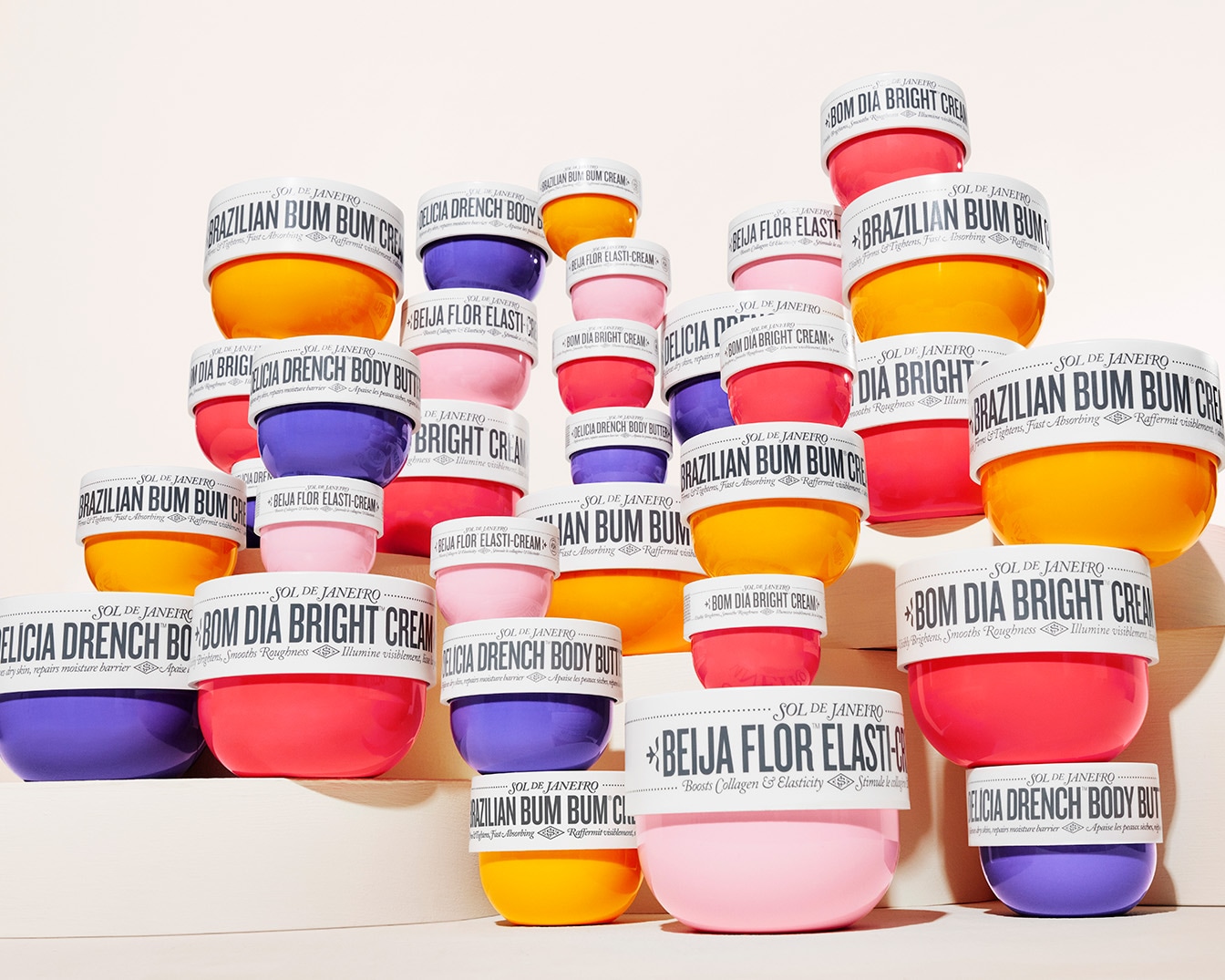 Descubre la colección Sol de Janeiro con cremas corporales en tonos vibrantes como amarillo, rosa, morado y naranja, cada una con una etiqueta blanca que detalla sus beneficios únicos.