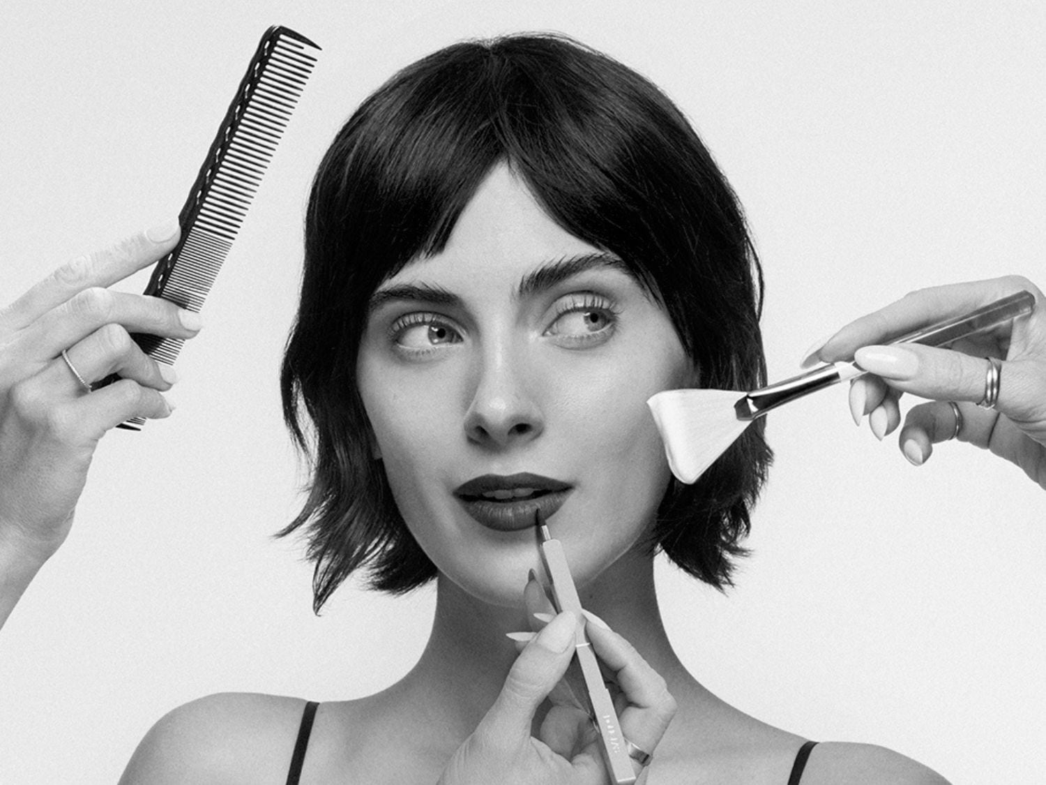 Čiernobiela fotografia ženy s krátkymi vlasmi, ktorej upravujú vlasy hrebeňom, nanášajú make-up štetcom a ceruzkou na pery.