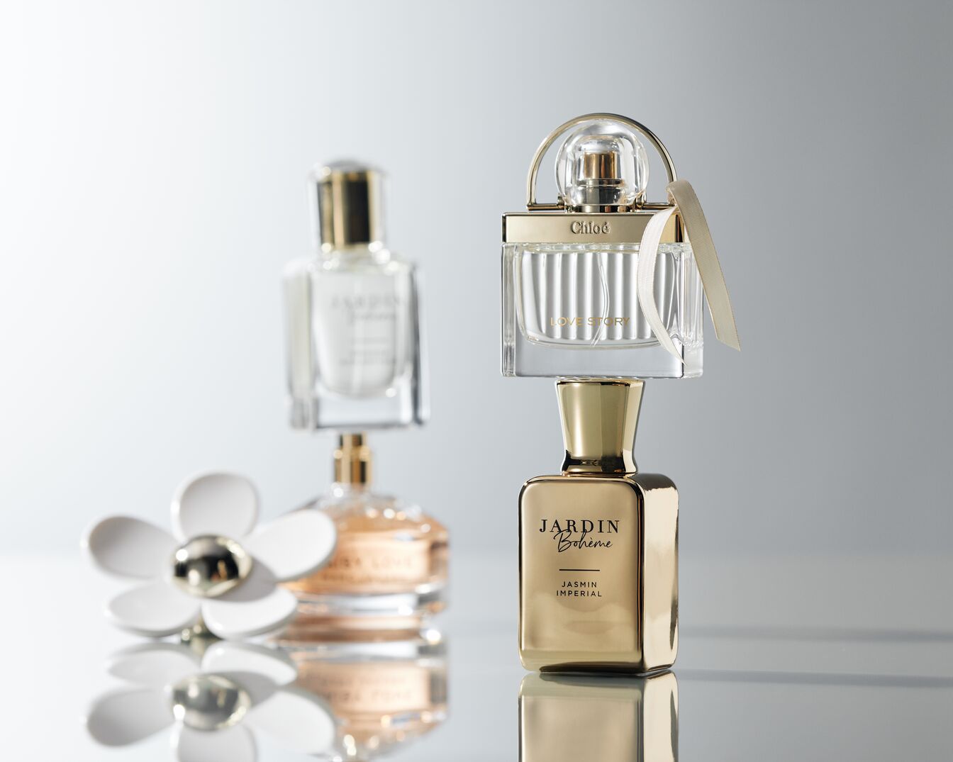 A képen három parfüm látható: egy arany Jardin Bohème Jasmin Imperial, egy Chloé Love Story és egy Marc Jacobs Daisy Love, egy fehér virág mellett.