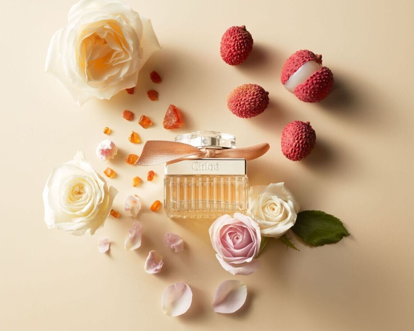 Chloé aromatas skaidraus stiklo buteliuke su kaspinu, apsuptas baltų ir rožinių rožių, ličių ir gintaro spalvos kristalų ant smėlio spalvos fono.