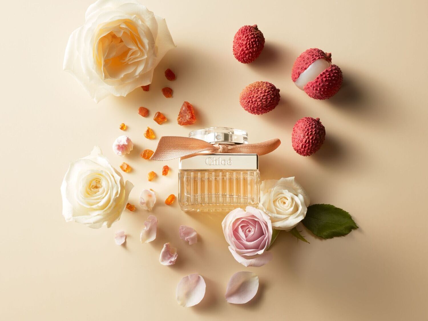 Chloé aromatas stikliniame buteliuke su rausvu kaspinu, apsuptas baltų rožių, ličių ir gintaro spalvos akmenukų ant smėlio spalvos fono.
