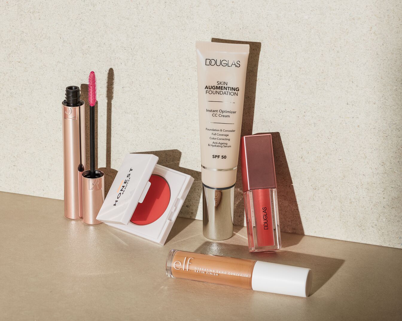 A Honest Beauty pirosító fehér tokban, egy Douglas Skin Augmenting Foundation CC krém SPF 50-nel, egy e.l.f. Hydrating Camo korrektor és egy Douglas szájfény látható.