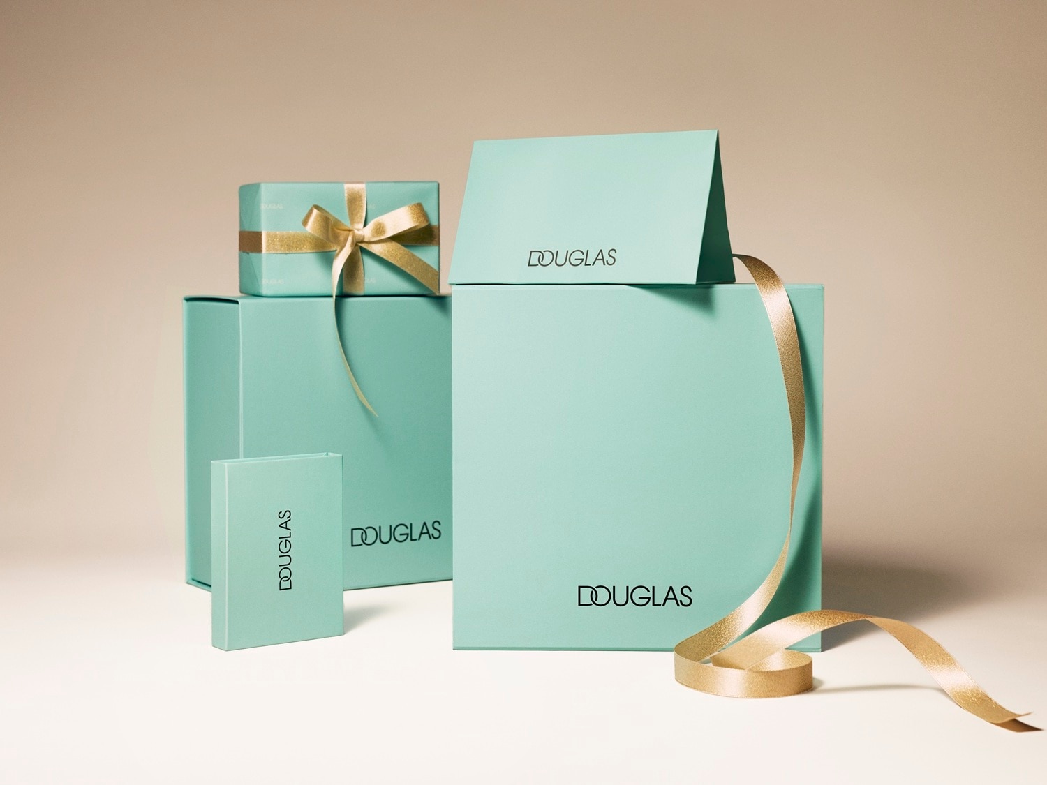 Na beżowym tle znajdują się turkusowe pudełka Douglas w różnych rozmiarach, przewiązane złotą wstążką, tworząc elegancką kompozycję prezentową.