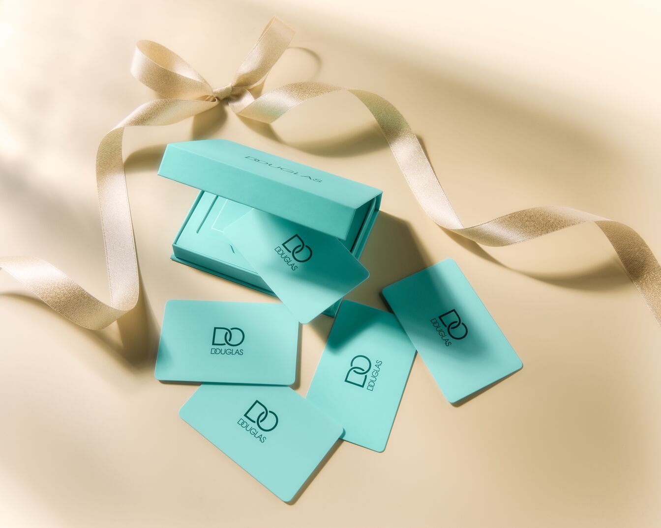 Na beżowym tle leżą turkusowe karty podarunkowe Douglas, obok nich znajduje się otwarte pudełko w tym samym kolorze i złota wstążka.