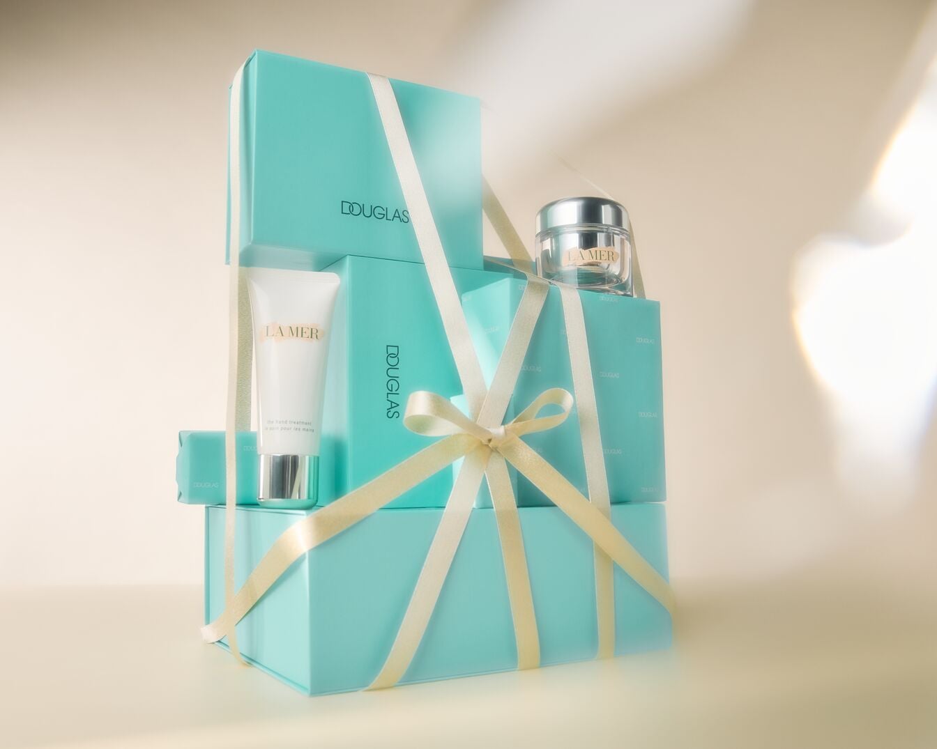 Ein Turm aus türkisfarbenen Douglas-Geschenkboxen, umwickelt mit cremefarbenen Bändern, präsentiert La Mer Hautpflegeprodukte: eine Tube Handcreme und ein Tiegel Gesichtscreme.