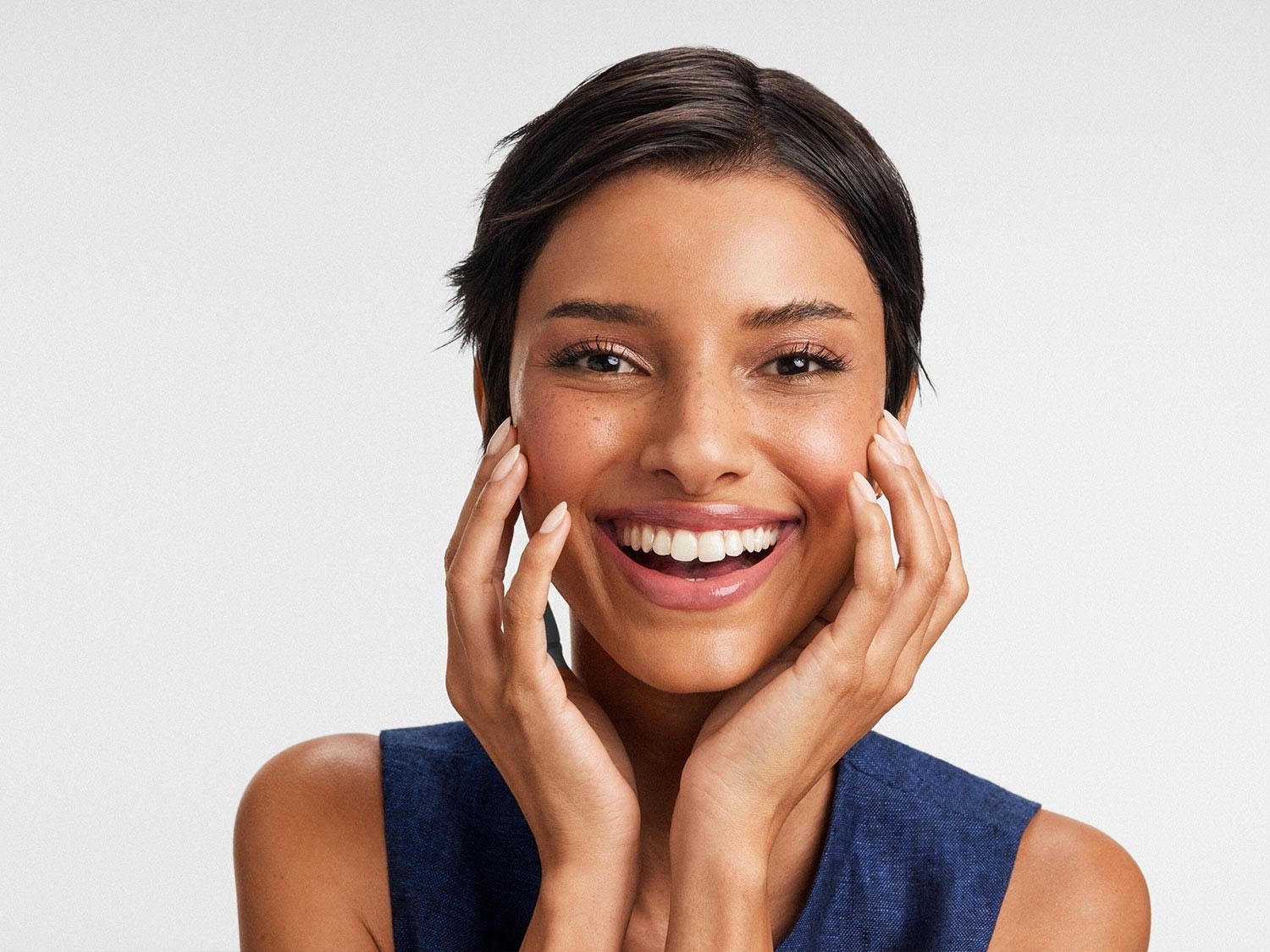 Une femme rayonnante avec un teint hâlé, les mains doucement posées sur ses joues, affichant un sourire éclatant qui illumine son visage.