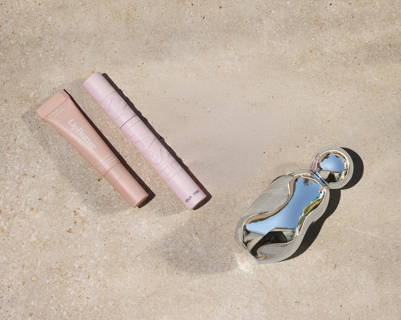 Ecco un trio di prodotti Kylie Skin: un Lip Butter color pesca in un tubetto, un mascara rosa chiaro e un profumo argentato dalla forma organica.