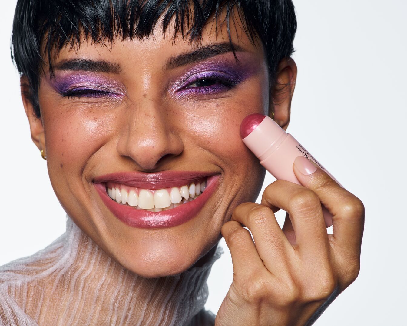 Uśmiechnięta kobieta z krótkimi, ciemnymi włosami i fioletowym makijażem oczu aplikuje róż w sztyfcie na policzek, trzymając go w dłoni.