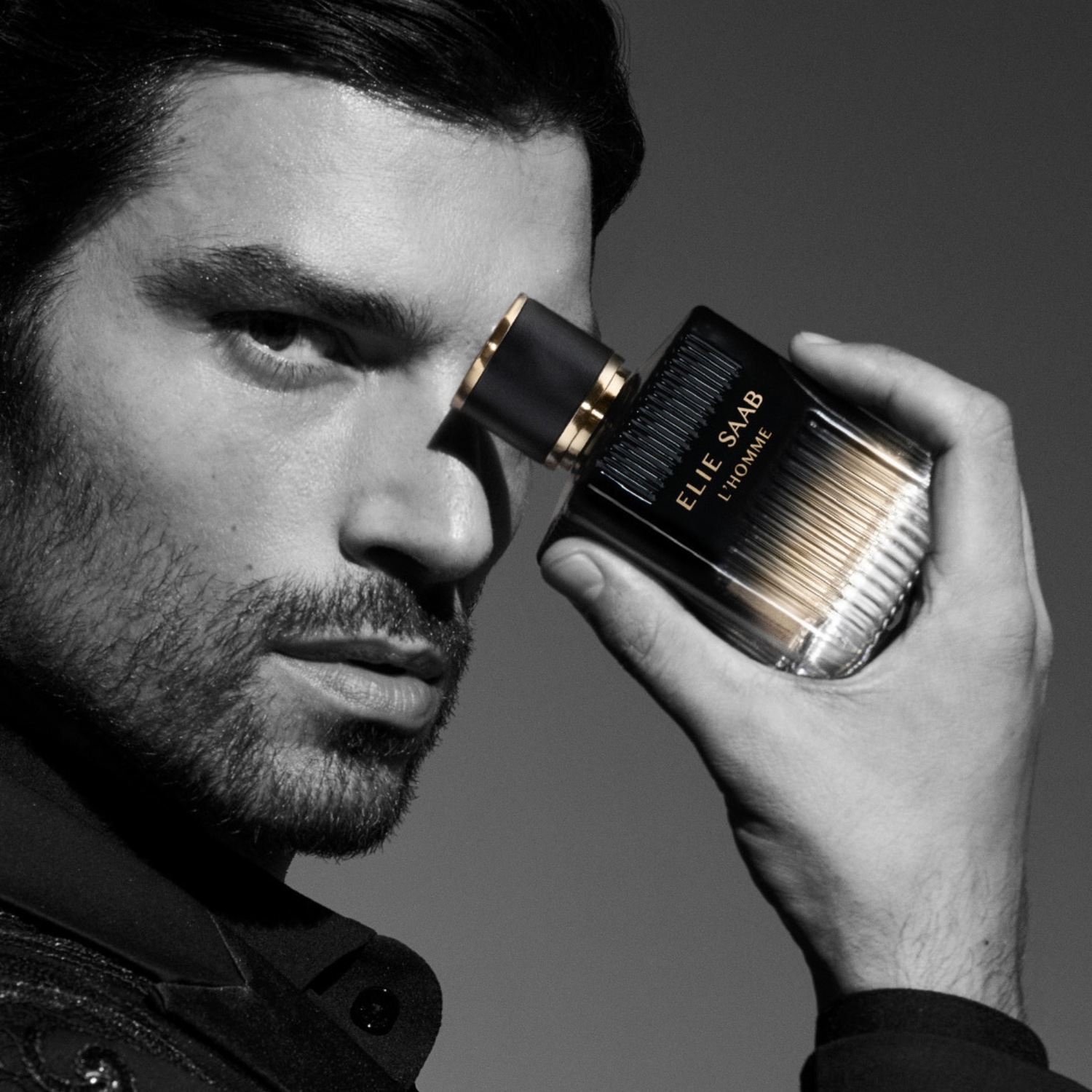 Czarno-białe zdjęcie mężczyzny trzymającego flakon perfum ELIE SAAB L'HOMME, z bliska widoczna jego twarz i dłoń.