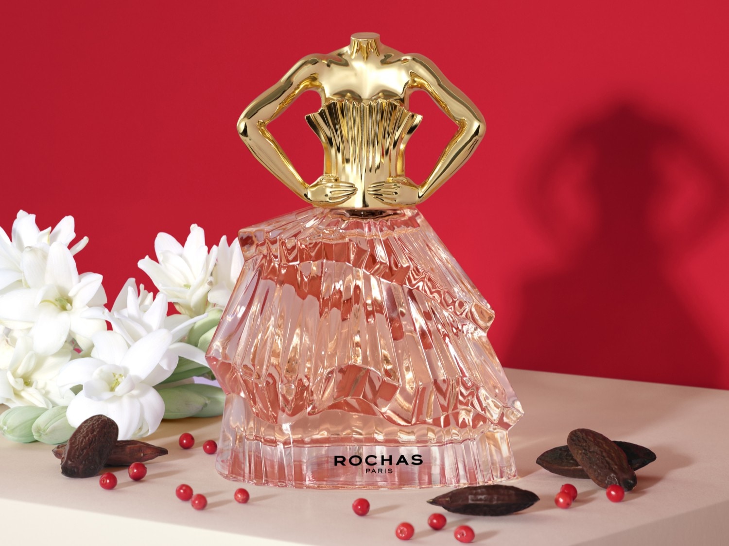 Elegancki flakon perfum Rochas Paris o kształcie sukni z pozłacanym gorsetem na tle czerwieni, otoczony białymi kwiatami i czerwonymi jagodami.