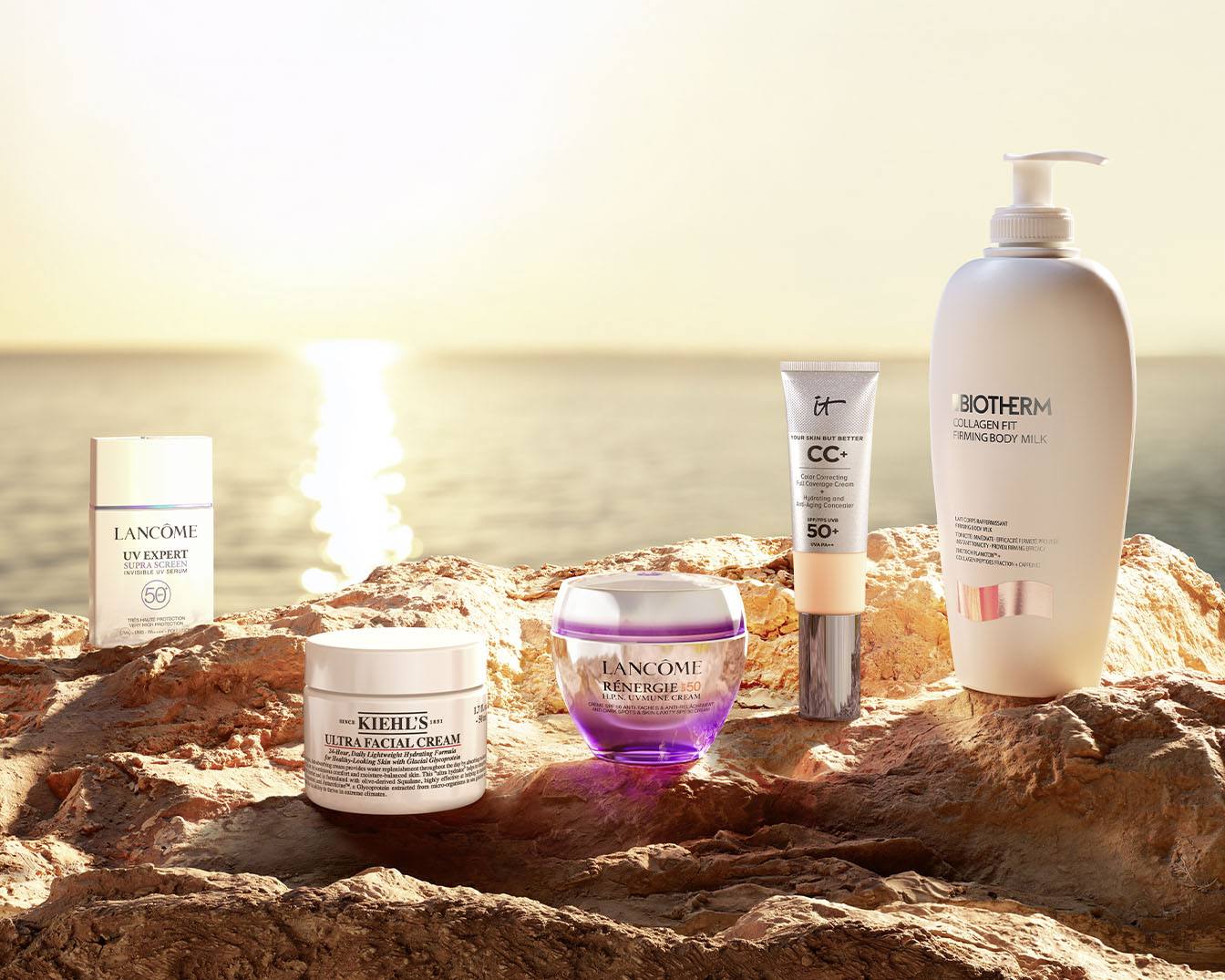 Eine Auswahl an Hautpflegeprodukten, darunter Lancôme UV Expert Serum, Kiehl's Ultra Facial Cream, Lancôme Rénergie Creme, IT CC+ Cream und Biotherm Collagen Fit Body Milk, auf einem Felsen mit Meerblick.