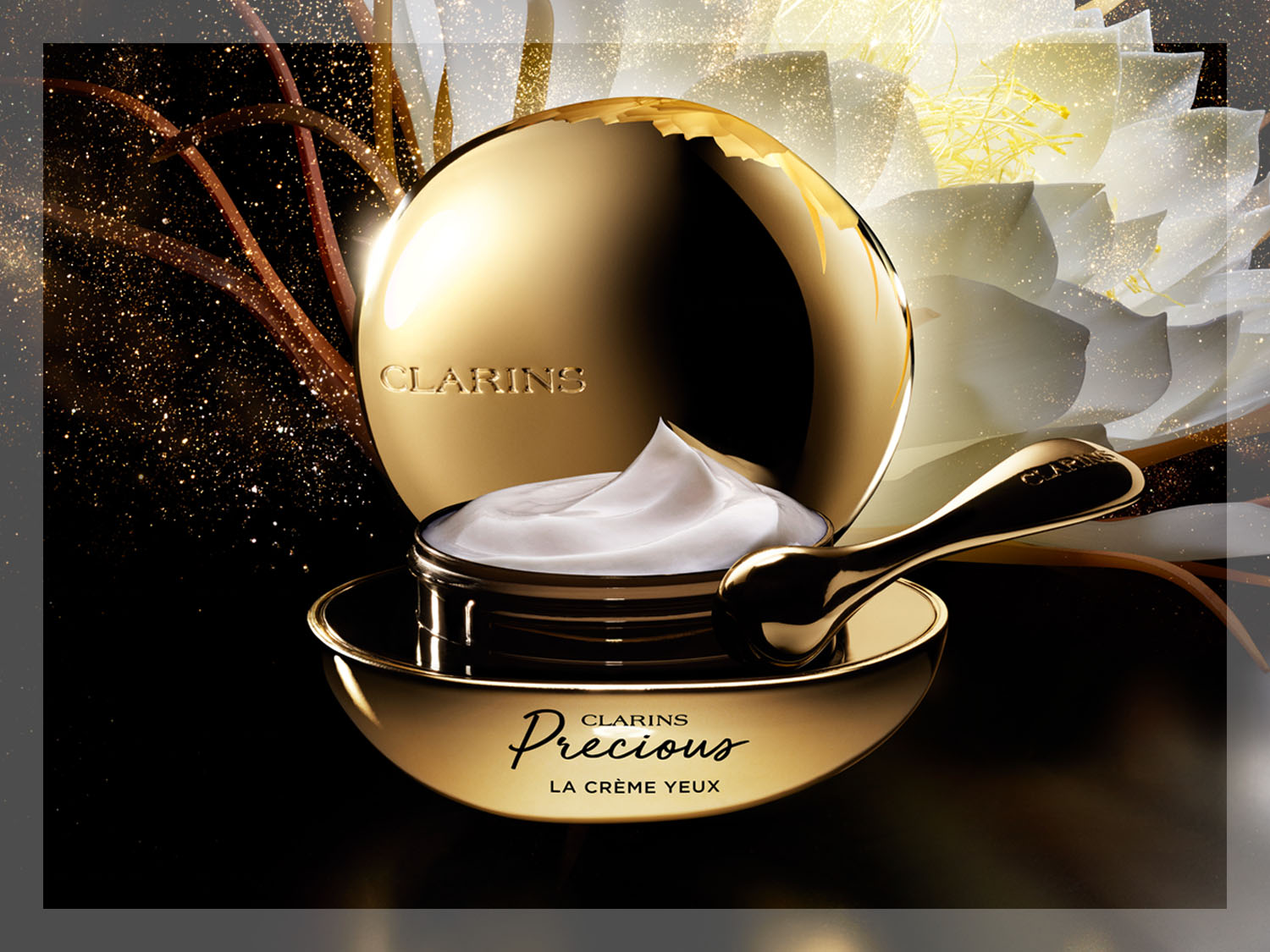 Złoty słoiczek Clarins Precious La Crème Yeux z białą zawartością i złotą szpatułką na tle kwiatów.