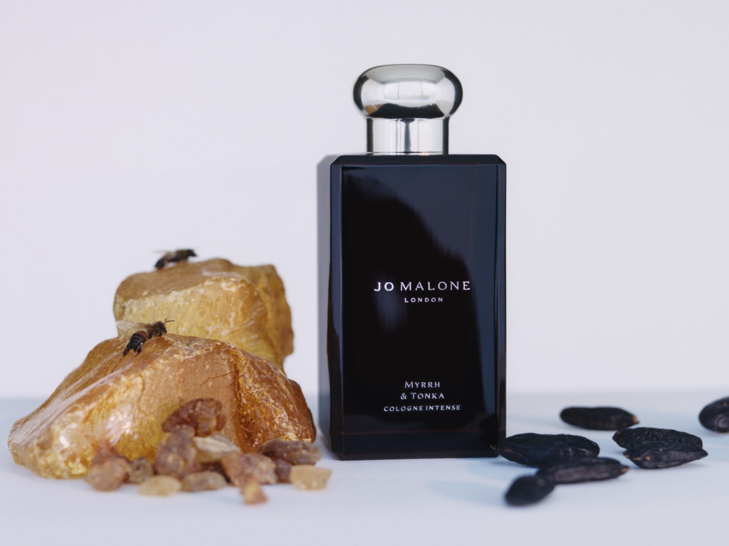 Czarny flakon Jo Malone London Myrrh & Tonka Cologne Intense stoi obok bryłek bursztynu z dwoma pszczołami i kilku czarnych ziaren.