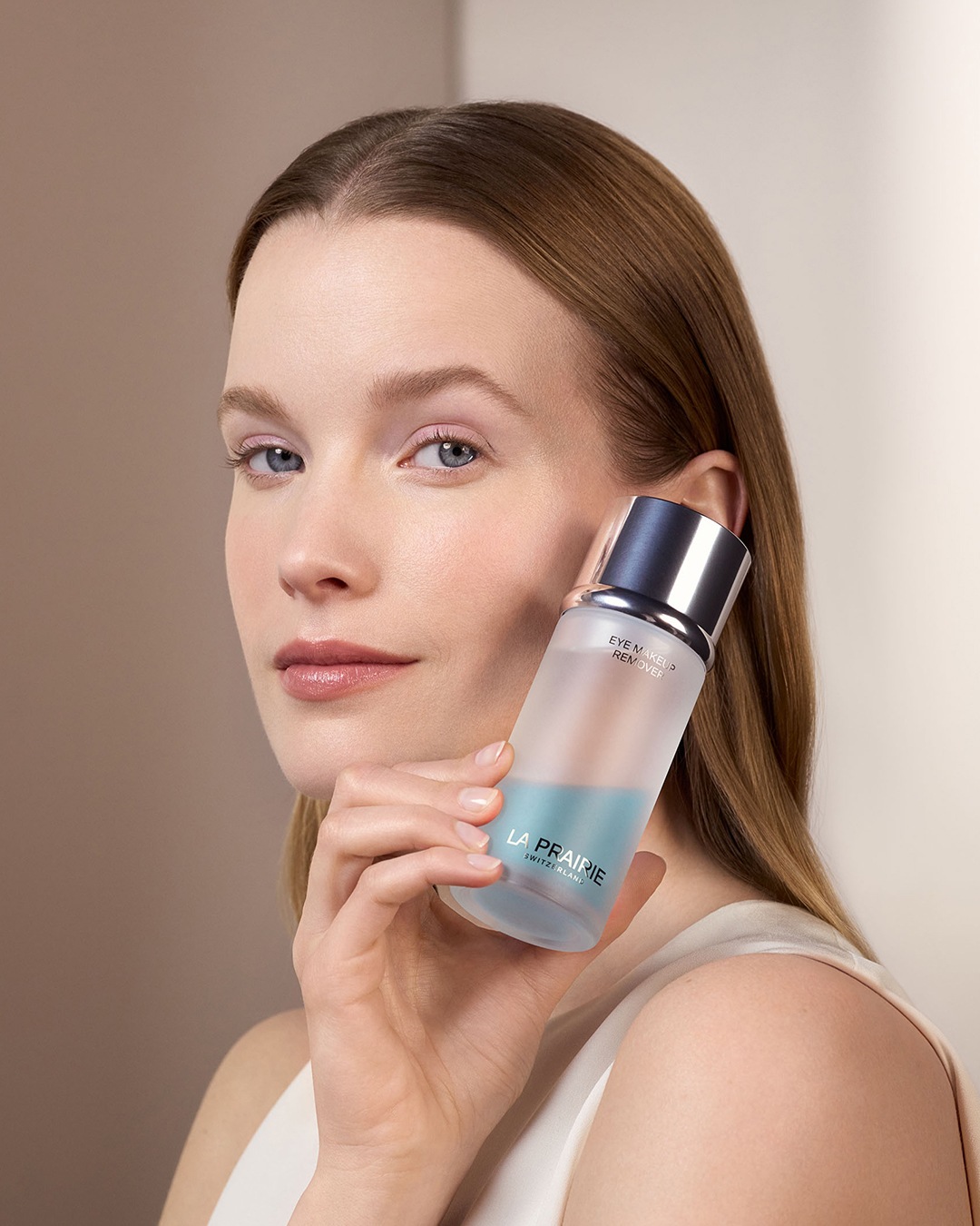 Une femme tient un flacon de démaquillant La Prairie, moitié transparent, moitié bleu, avec un bouchon argenté, pour un nettoyage doux et efficace des yeux.