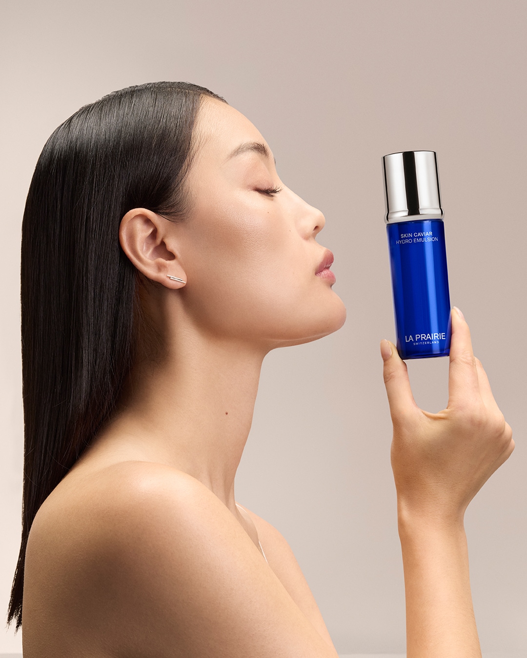 Une femme aux cheveux noirs lisses tient un flacon bleu de Skin Caviar Hydro Emulsion de La Prairie, les yeux fermés, savourant le parfum.