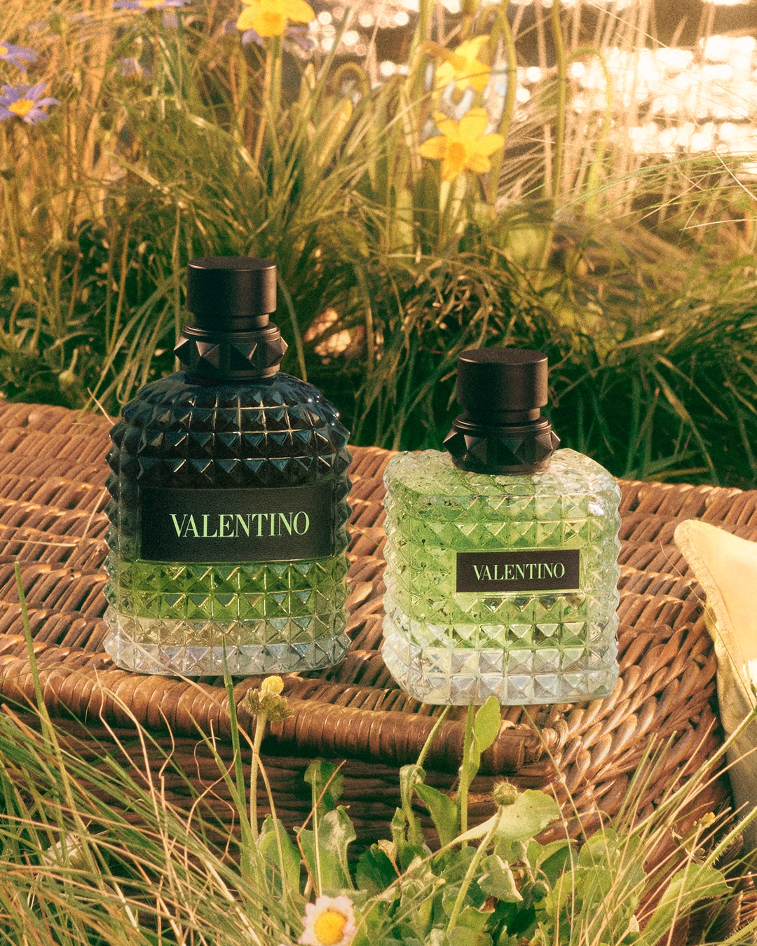 Deux flacons de parfum Valentino, l'un bleu foncé et l'autre vert clair, reposent sur un panier en osier dans un cadre champêtre.