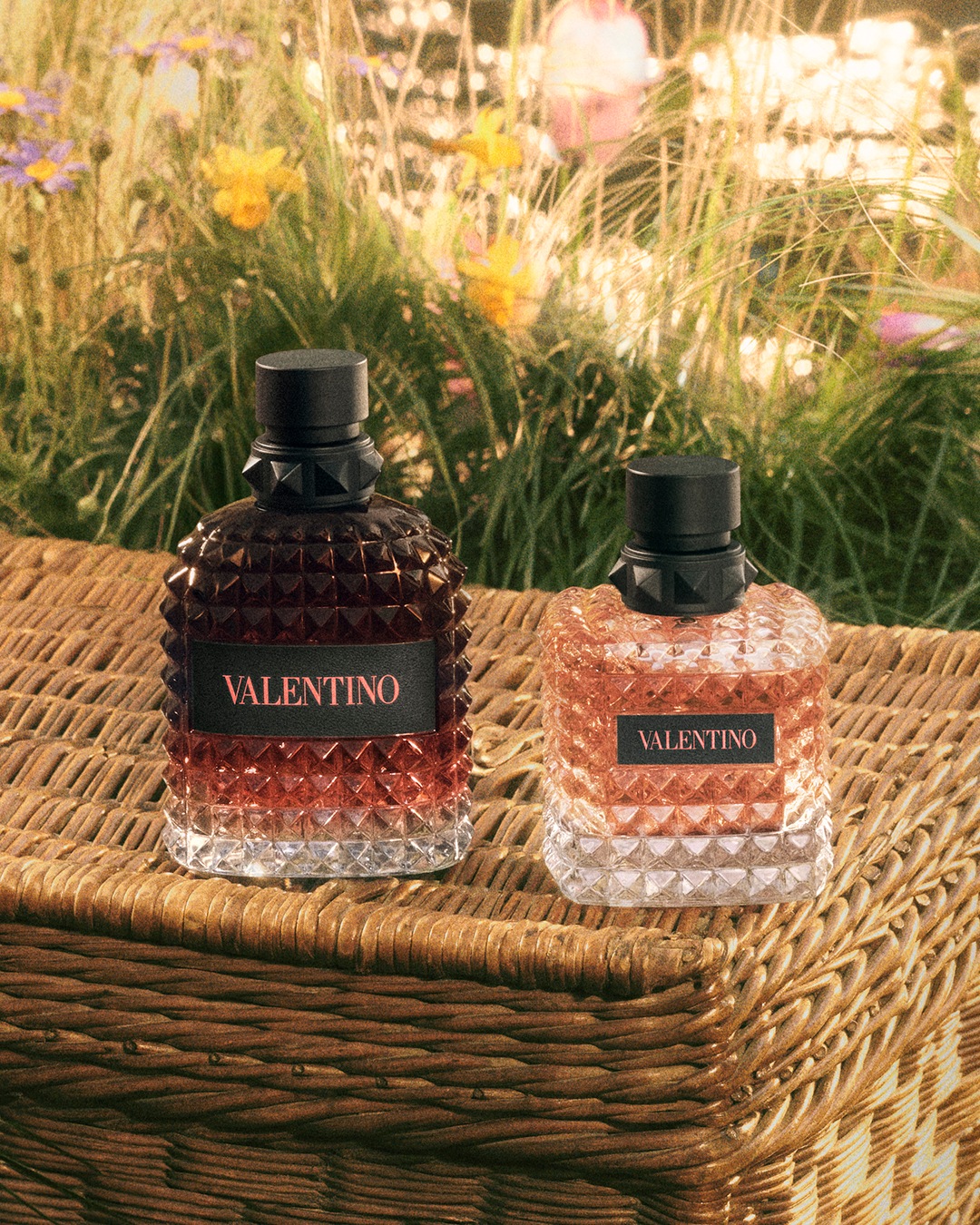 Deux flacons de parfum Valentino, l'un rouge foncé et l'autre rose clair, reposent sur un panier en osier tressé, avec un arrière-plan flou de fleurs sauvages et d'herbes hautes.