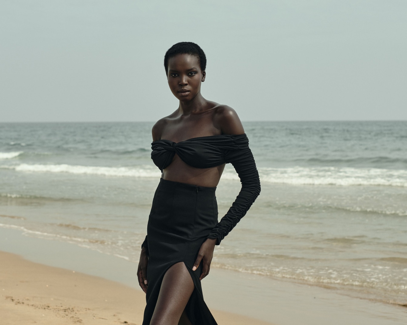 Na tle oceanu stoi modelka w czarnym dwuczęściowym stroju z odkrytymi ramionami i spódnicą z rozcięciem, prezentując elegancję i styl.